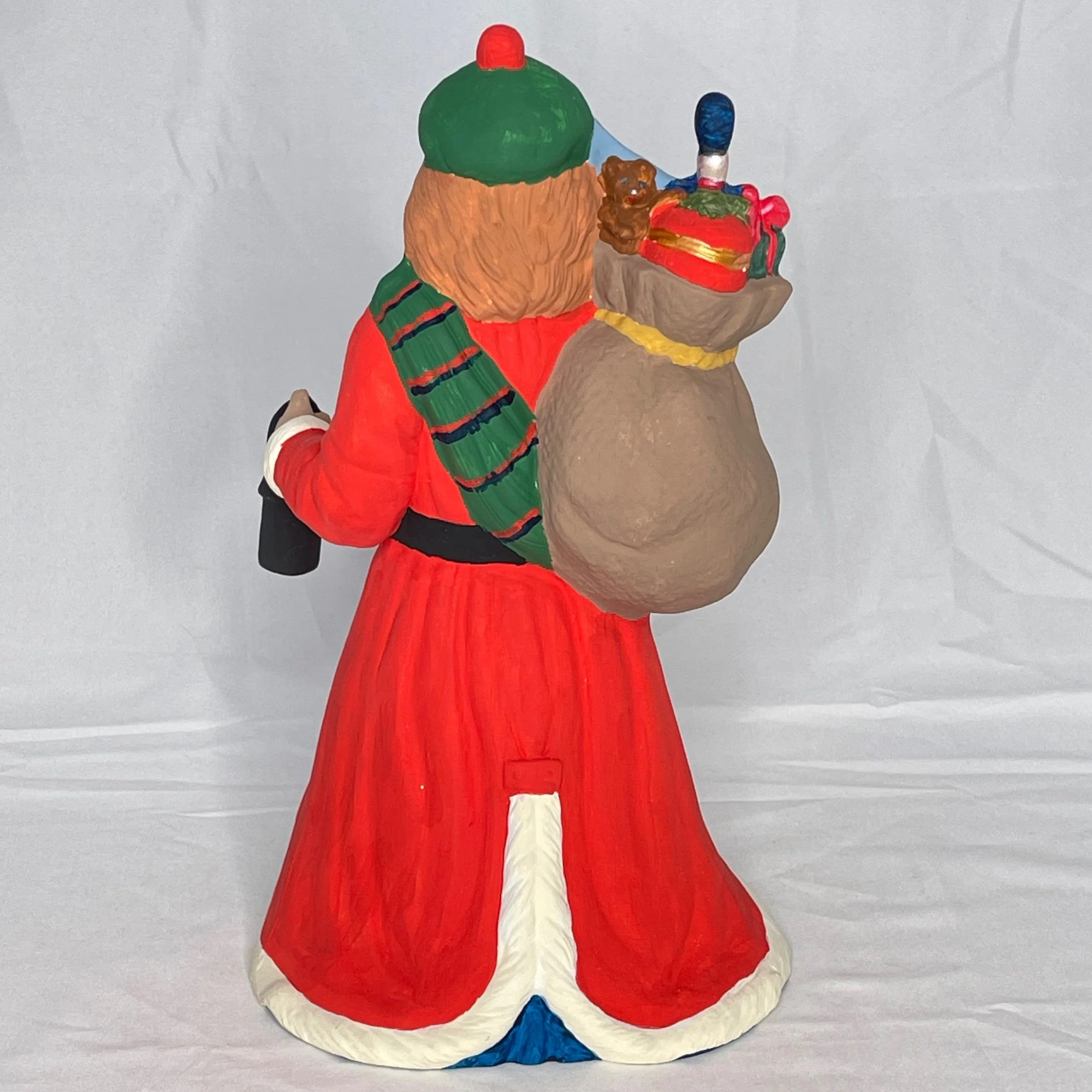 Hand-painted Christmas figurine of Santa Claus holding a bag of gifts, with a teddy bear and a toy soldier inside the bag, standing against a white background.