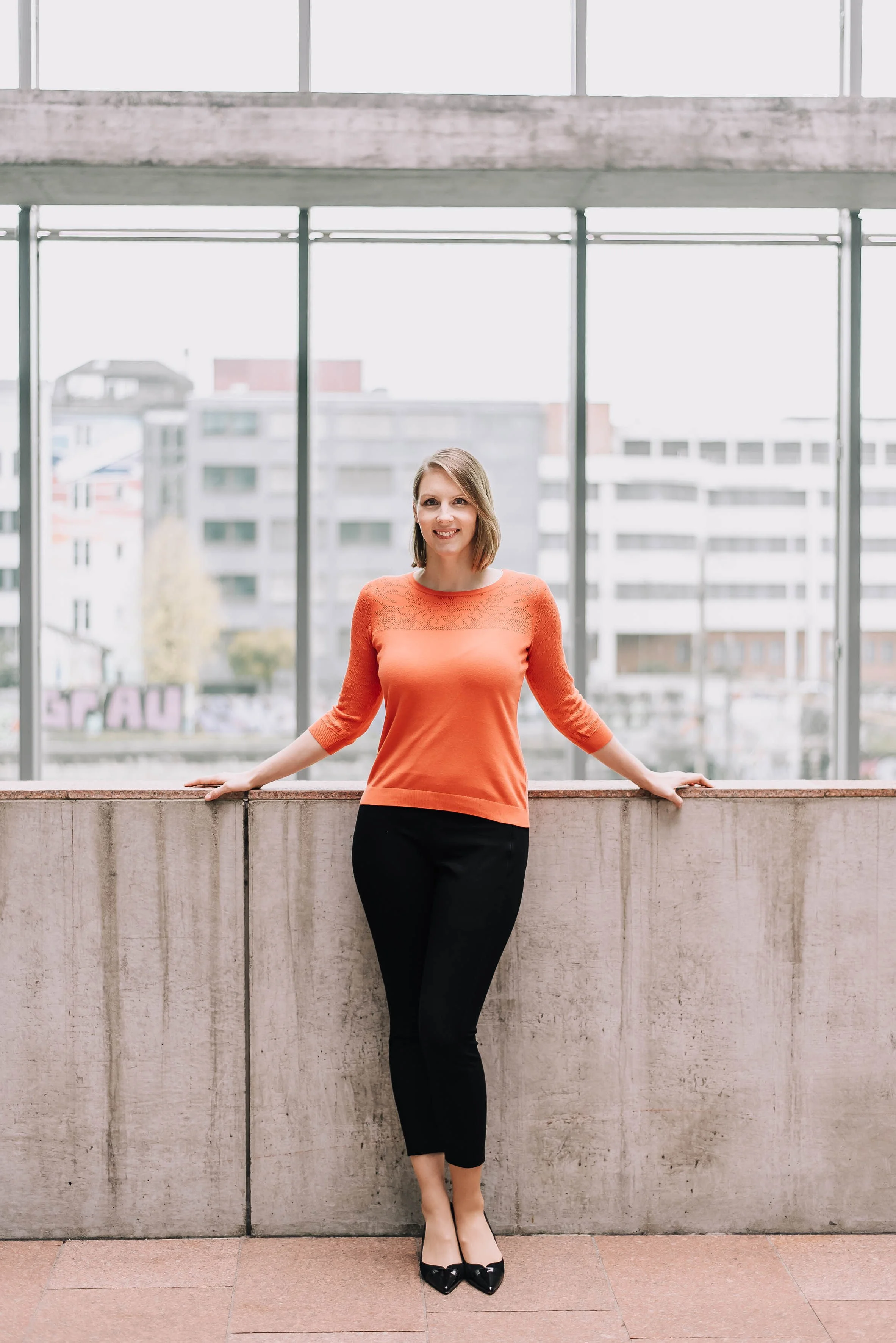 Junge Frau steht vor einer Betonbrüstung in einem modernen Innenraum mit großen Fenstern und einer städtischen Skyline im Hintergrund.