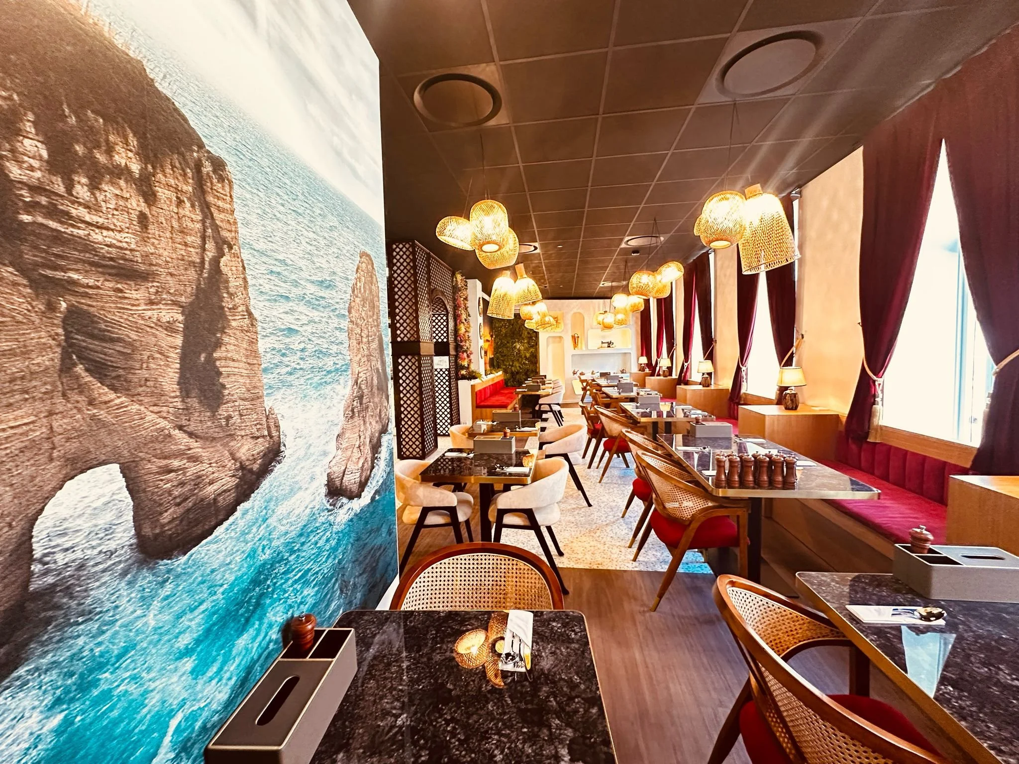 Interior of a modern restaurant with a large ocean mural on the wall, dark ceiling, hanging lights, and tables with red chairs and bench seating.