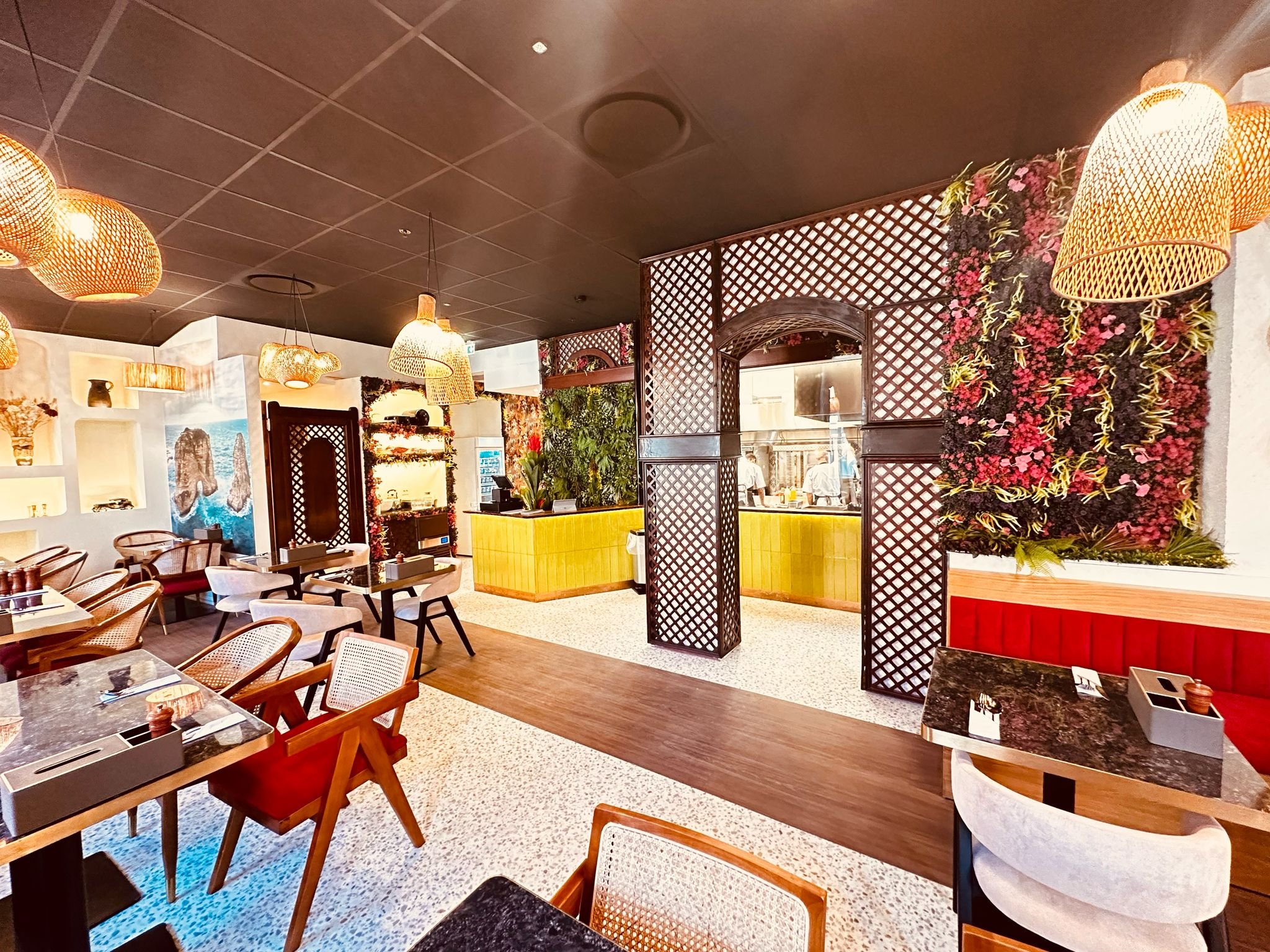 Interior of a modern restaurant with wooden and wicker furniture, decorative wall panels, and vibrant floral arrangements. The dining area features a mix of tables and chairs with a visible open kitchen area. Warm lighting adds to the cozy atmosphere