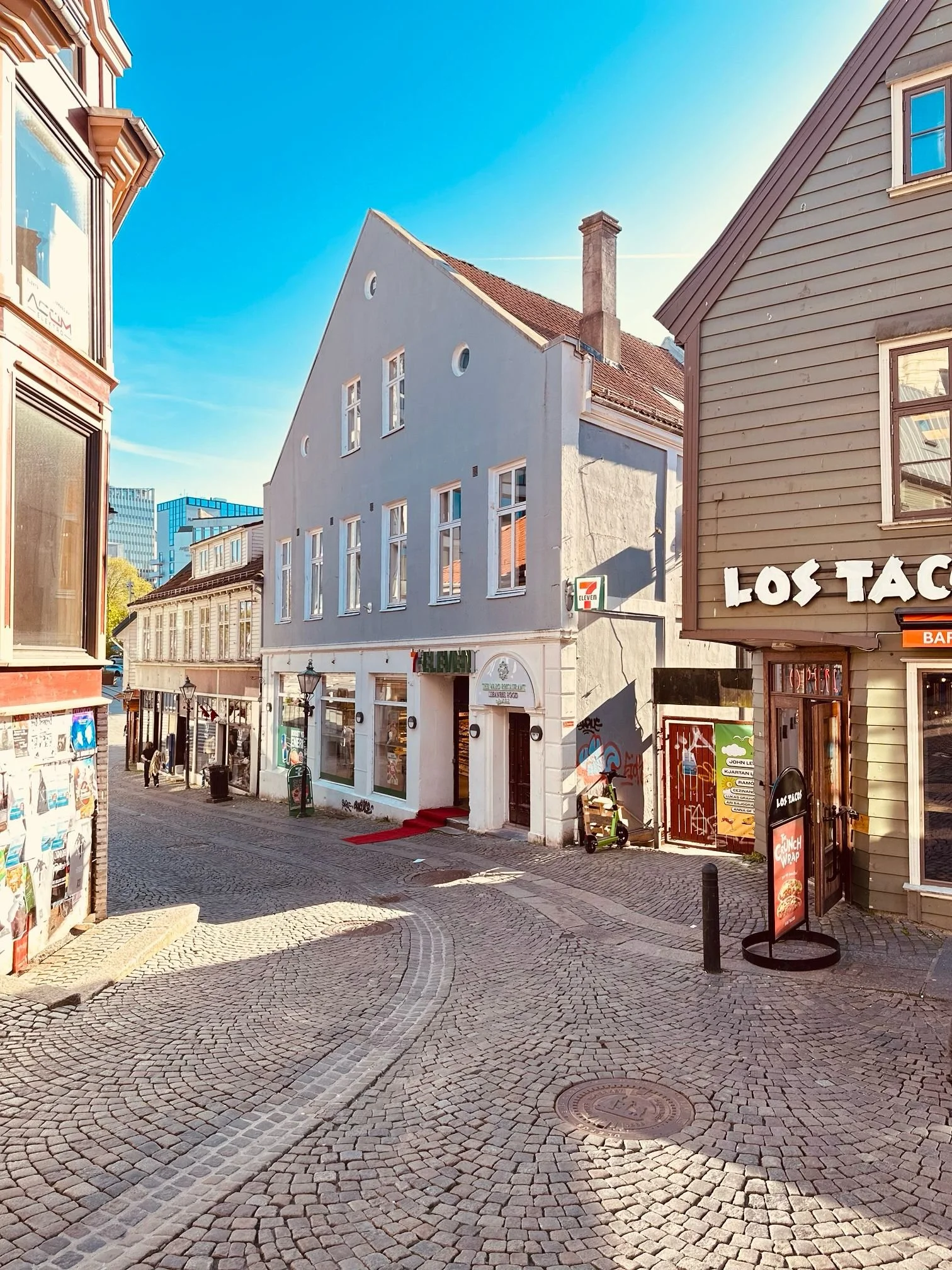 A quaint cobblestone street in a European city with colorful buildings, including a blue house and a restaurant named "Los Tacos." The street is lined with various signs and posters, and a 7-Eleven store is visible. A clear blue sky overhead.
