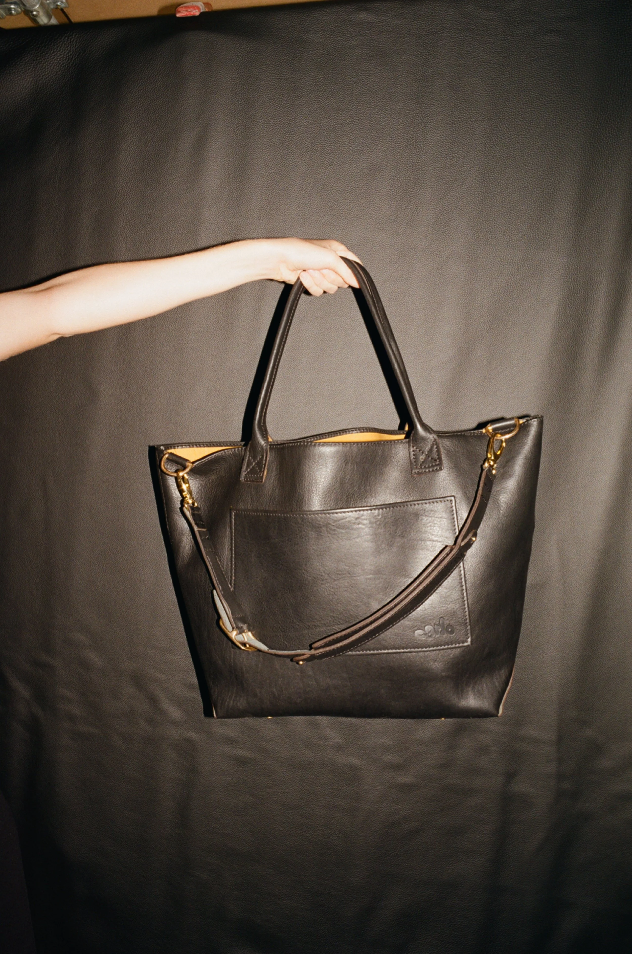 A black leather tote bag with shoulder straps is being held up by a person's arm against a dark background.