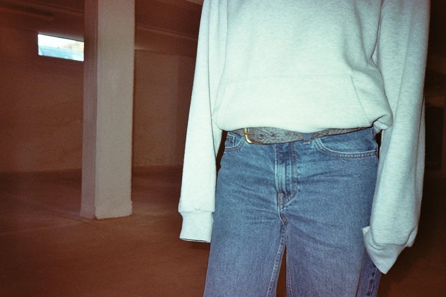 Close-up of a person wearing a light gray sweatshirt, blue jeans, and a patterned belt, standing in a room with wooden floors and white walls.