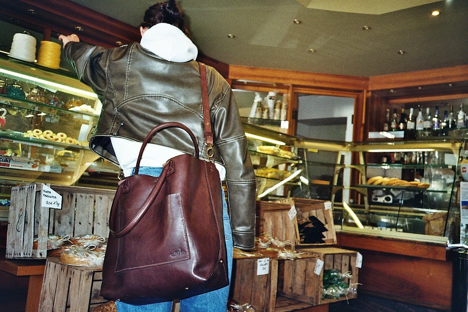 Person shopping at a bakery or deli counter with baked goods and bottles in the background, carrying a large brown leather tote bag.