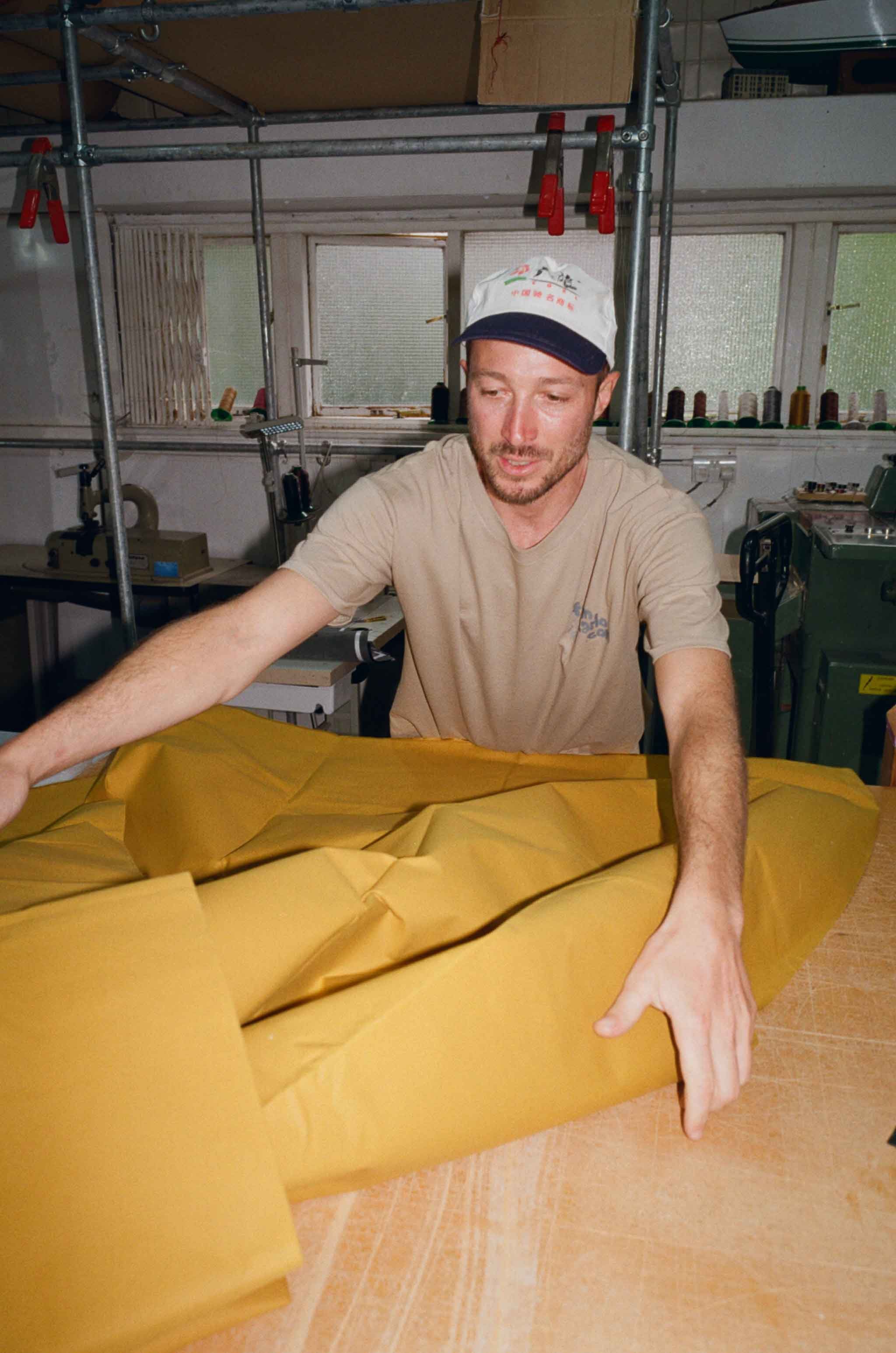 A man in a beige t-shirt and a white cap is folding bright yellow fabric at a workbench in a workshop with spools of thread and equipment in the background.