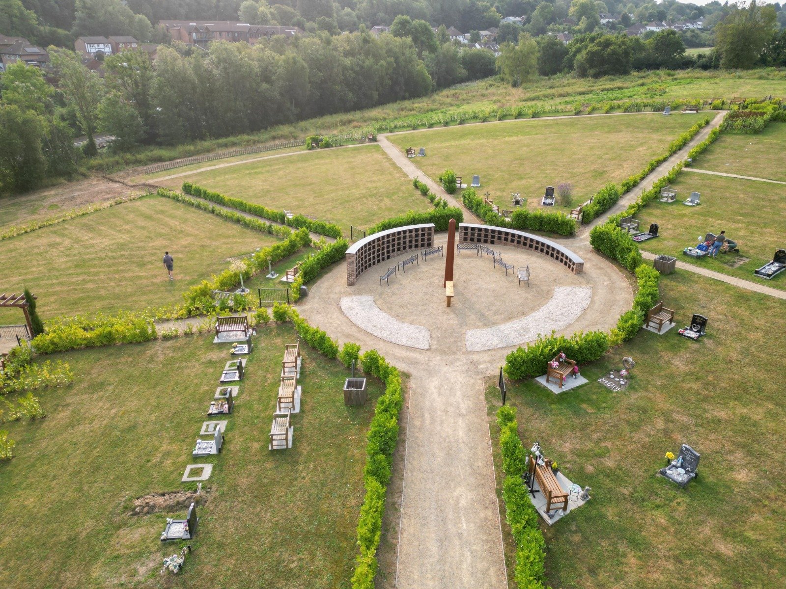 Aerial view of a cemetery with a circular path and benches, surrounded by green grass and trees. Gravestones are visible in rows, and a person is walking on a path. Residential buildings are in the background.