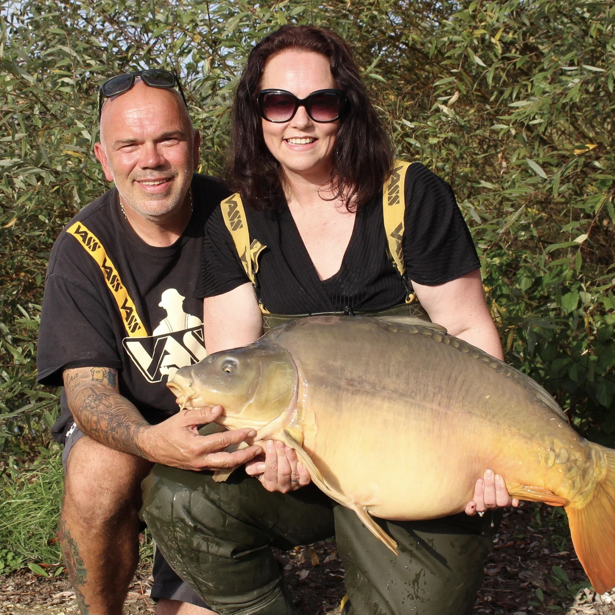 A man and a woman smiling outdoors, holding a large orange and gray fish together, surrounded by green bushes.