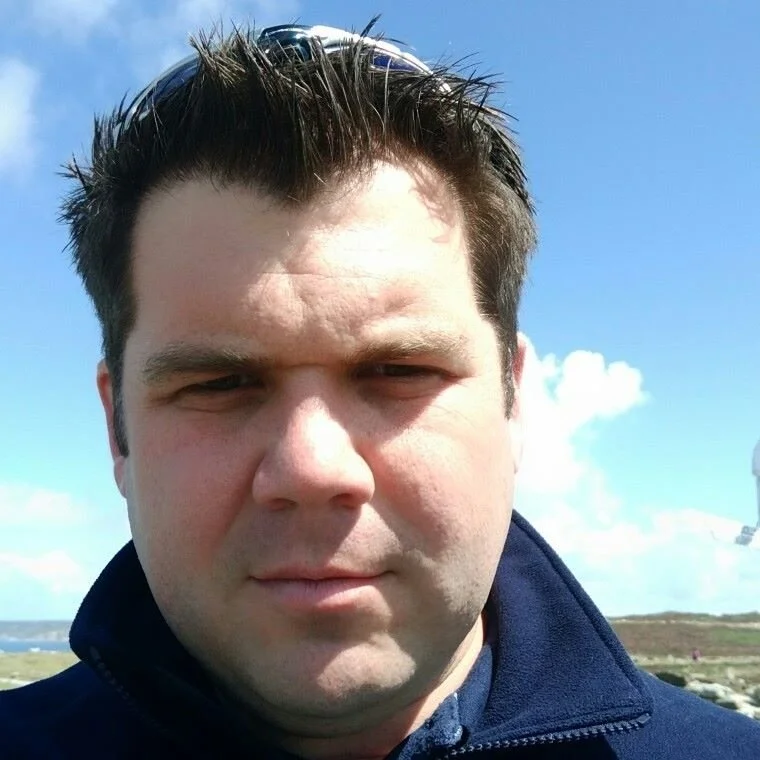 Close-up of a man with dark hair, wearing a dark jacket, outdoors on a sunny day with blue sky and clouds in the background.