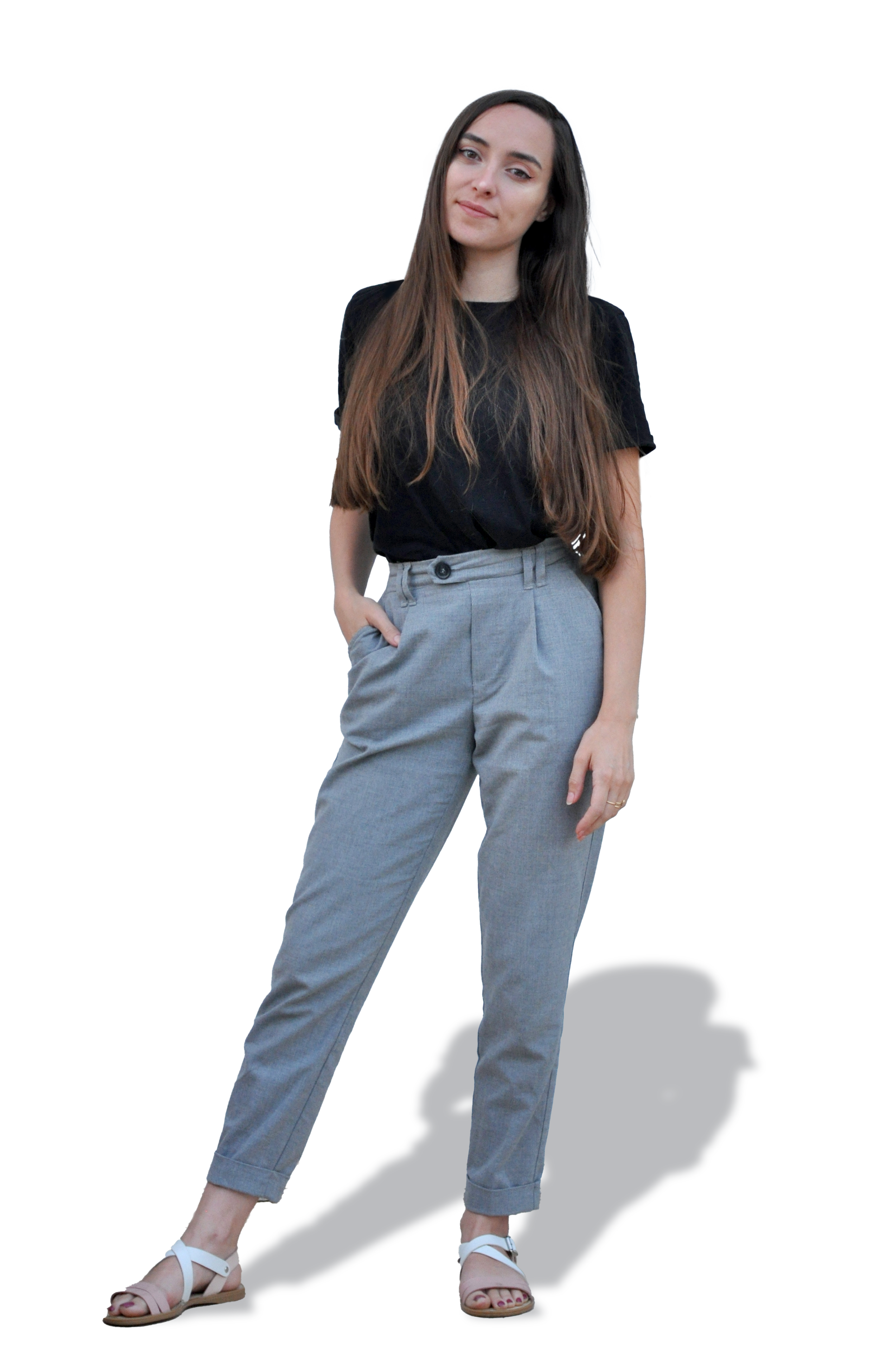 A woman with long brown hair wearing a black t-shirt, gray high-waisted pants, and white sandals standing with one hand in her pocket against a background with a large green rectangle and a colorful pattern.