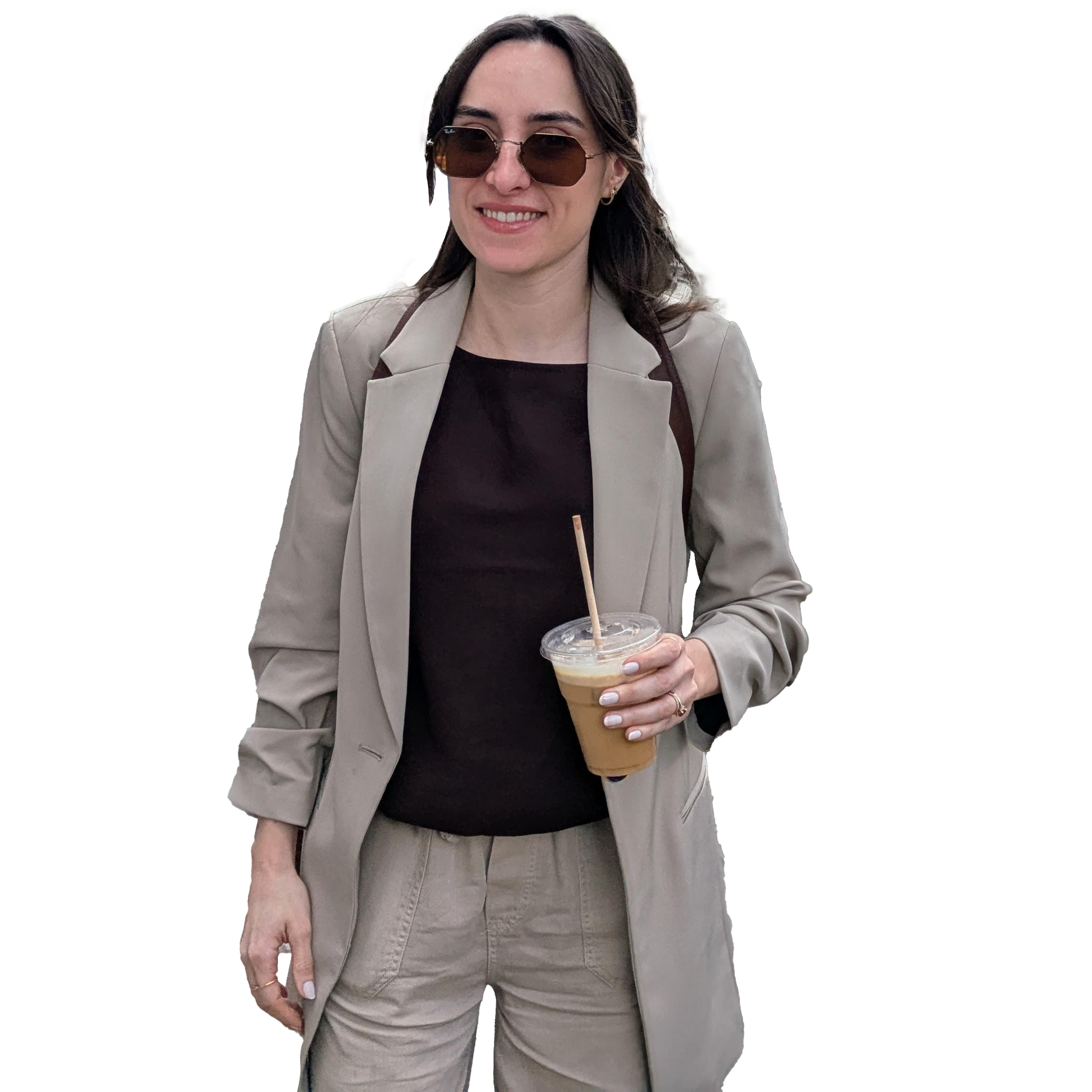 A woman with dark hair and sunglasses holding a cup of iced coffee, dressed in a beige suit with a black shirt, smiling.