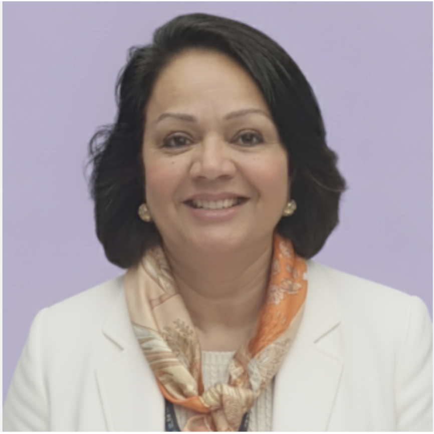 A woman with dark, shoulder-length hair smiling, wearing a white blazer, an orange patterned scarf, and pearl earrings, standing against a light purple background.