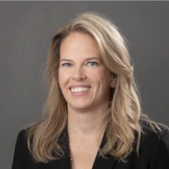 A woman who specializes in genius children with high IQs with long, wavy blonde hair wearing a black blazer smiling against a gray background.