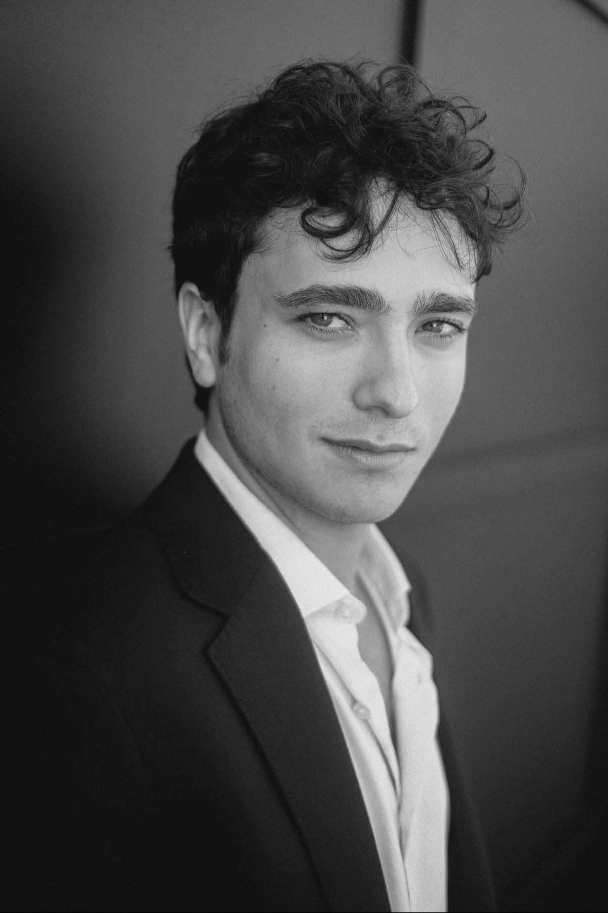 Black and white portrait of a young famous superhero actor with curly hair, wearing a dark blazer and white shirt, smiling slightly, looking into the camera.