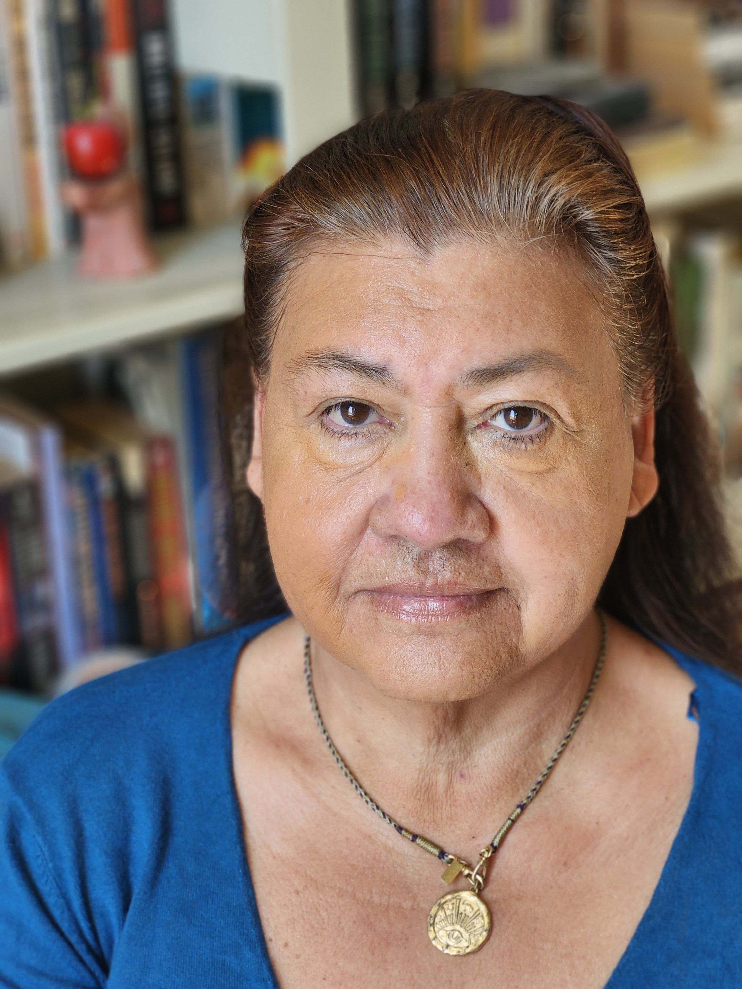 Close-up of a woman with brown hair and multiple necklaces, wearing a blue top, standing in front of a bookshelf.