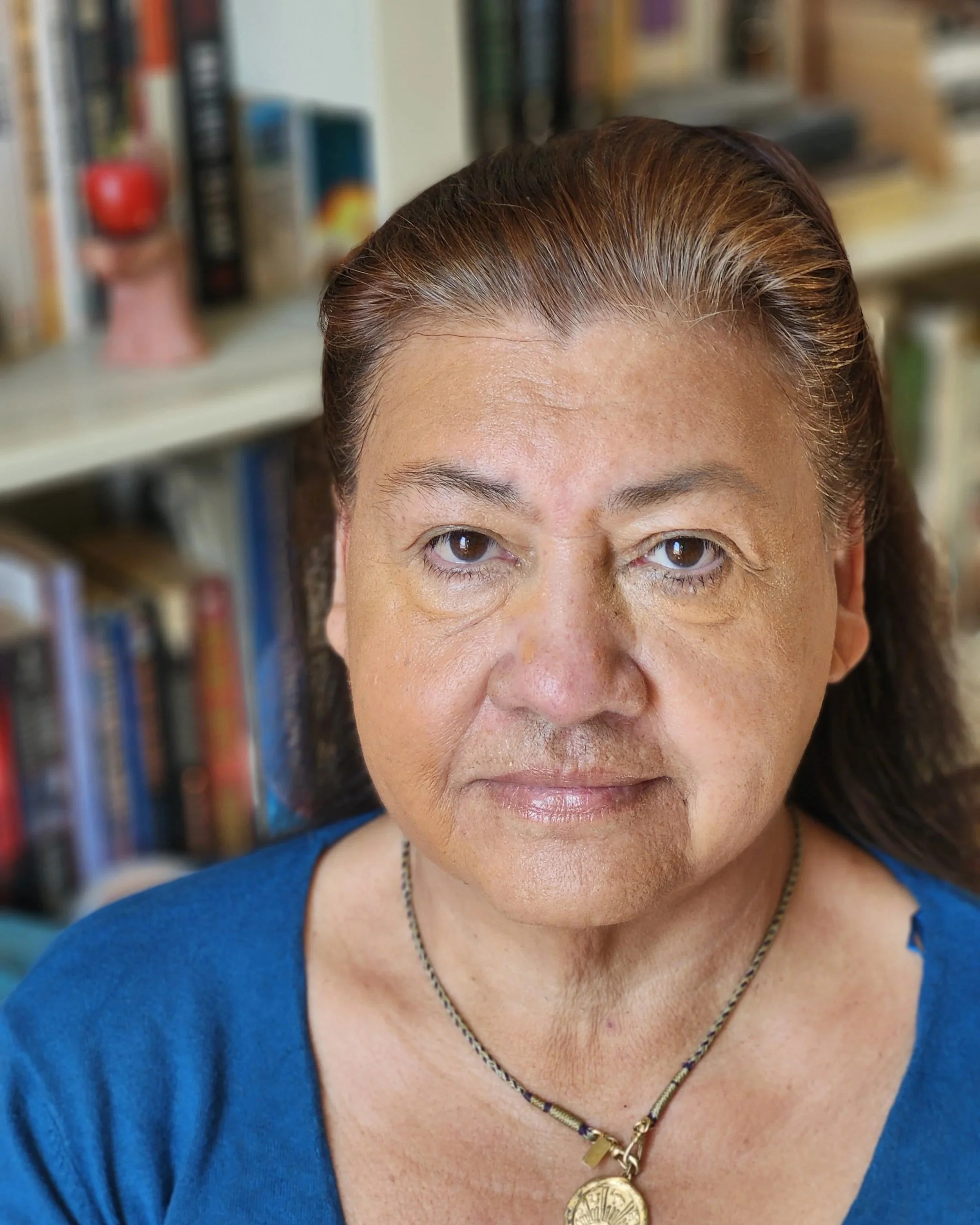 Close-up of a migrant expert with brown hair, wearing a blue top and a gold necklace with religious medallion, in front of a bookshelf filled with books.