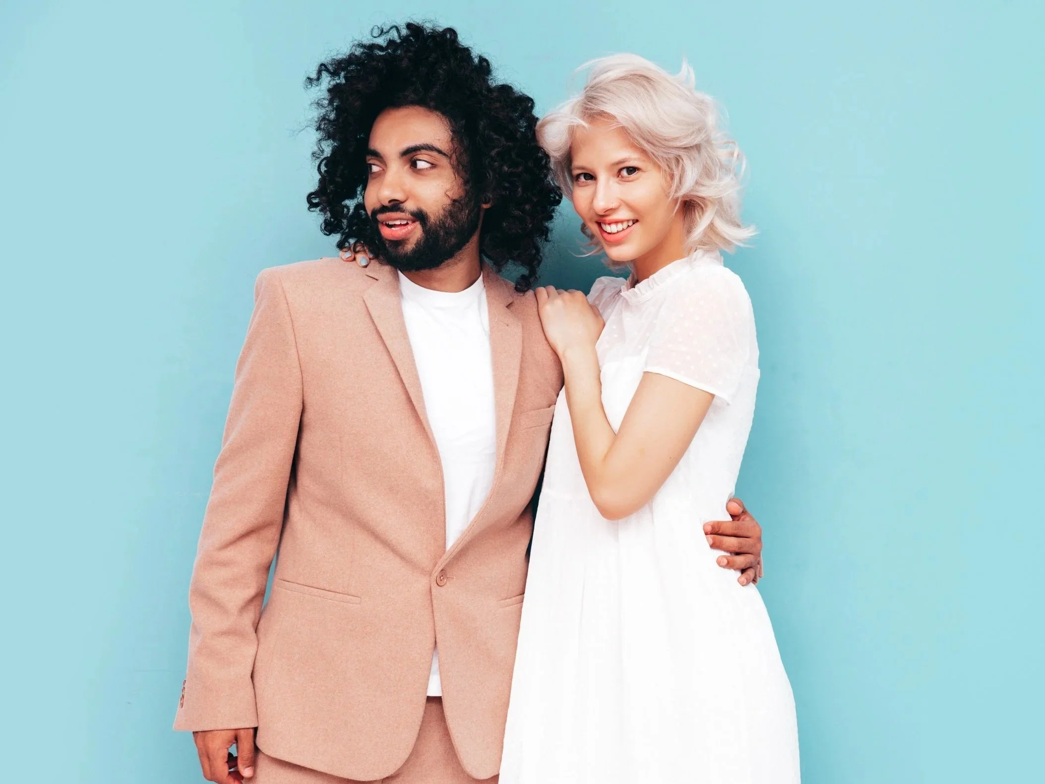 A young man with curly black hair and a beard wearing a beige suit and white shirt, standing next to a young woman with short platinum blonde hair wearing a white dress, against a light blue background.
