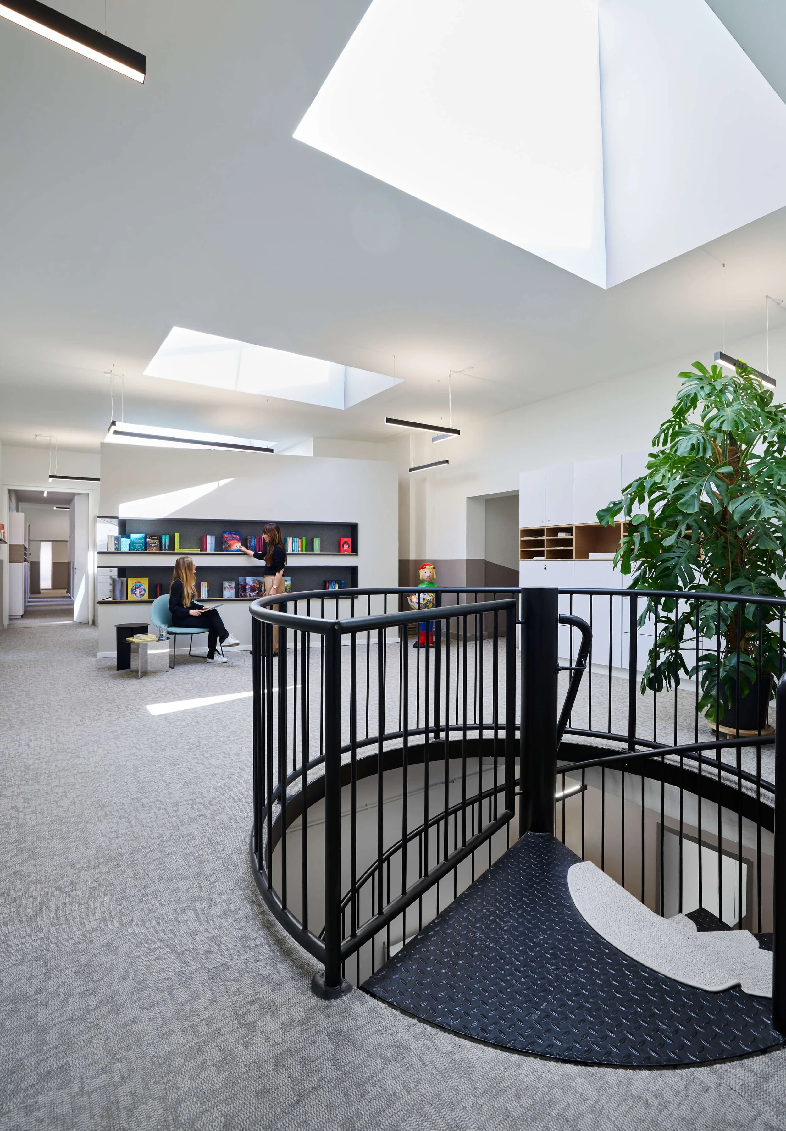 Heller Innenraum mit Wendeltreppe, Oberlichtern, Bücherregal, Pflanzen und zwei Personen im Sitzbereich.