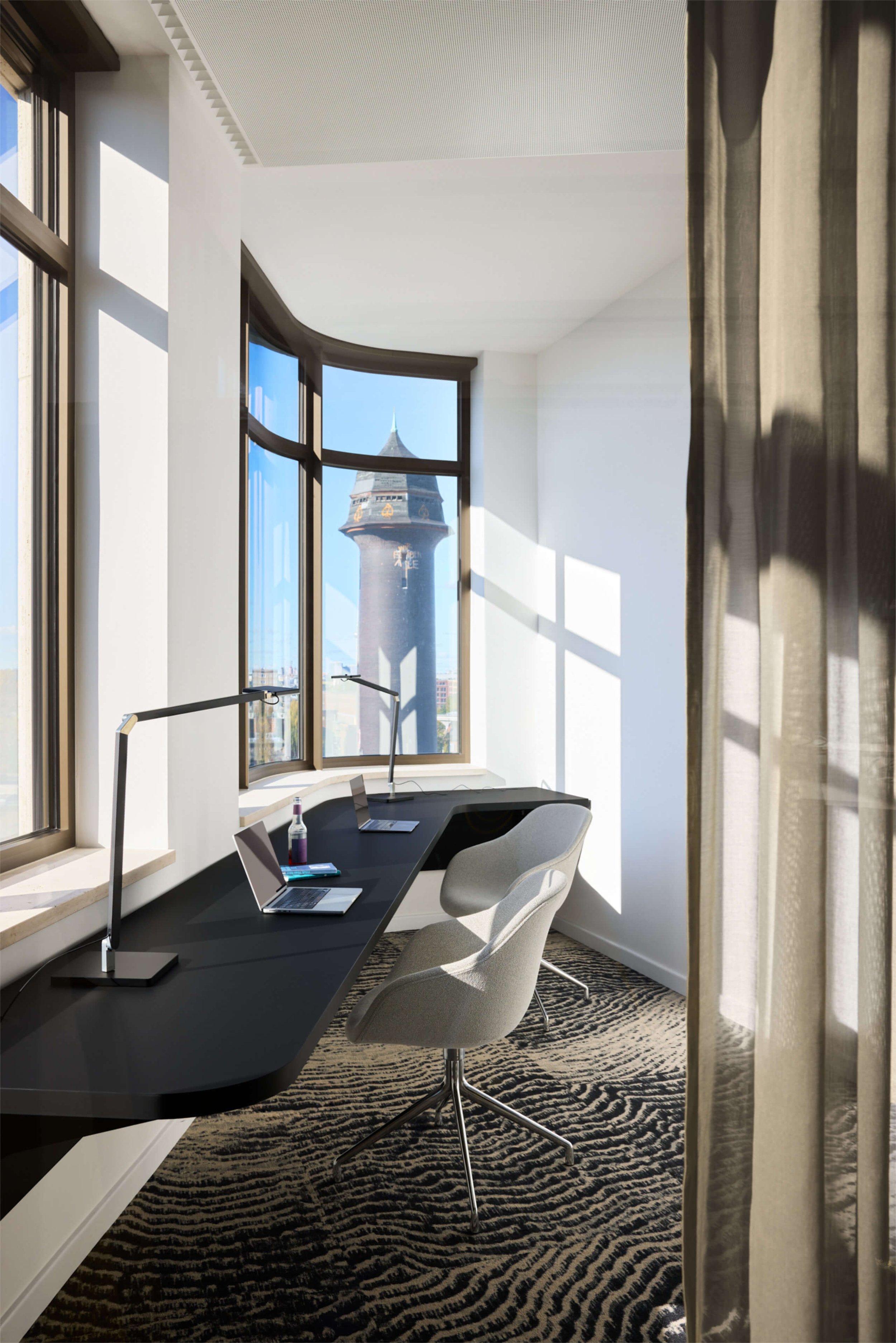 Arbeitsplatz an einer Fensterfront mit gebogenem Schreibtisch, Stuhl, Leuchten, Laptop und Blick auf einen Turm.