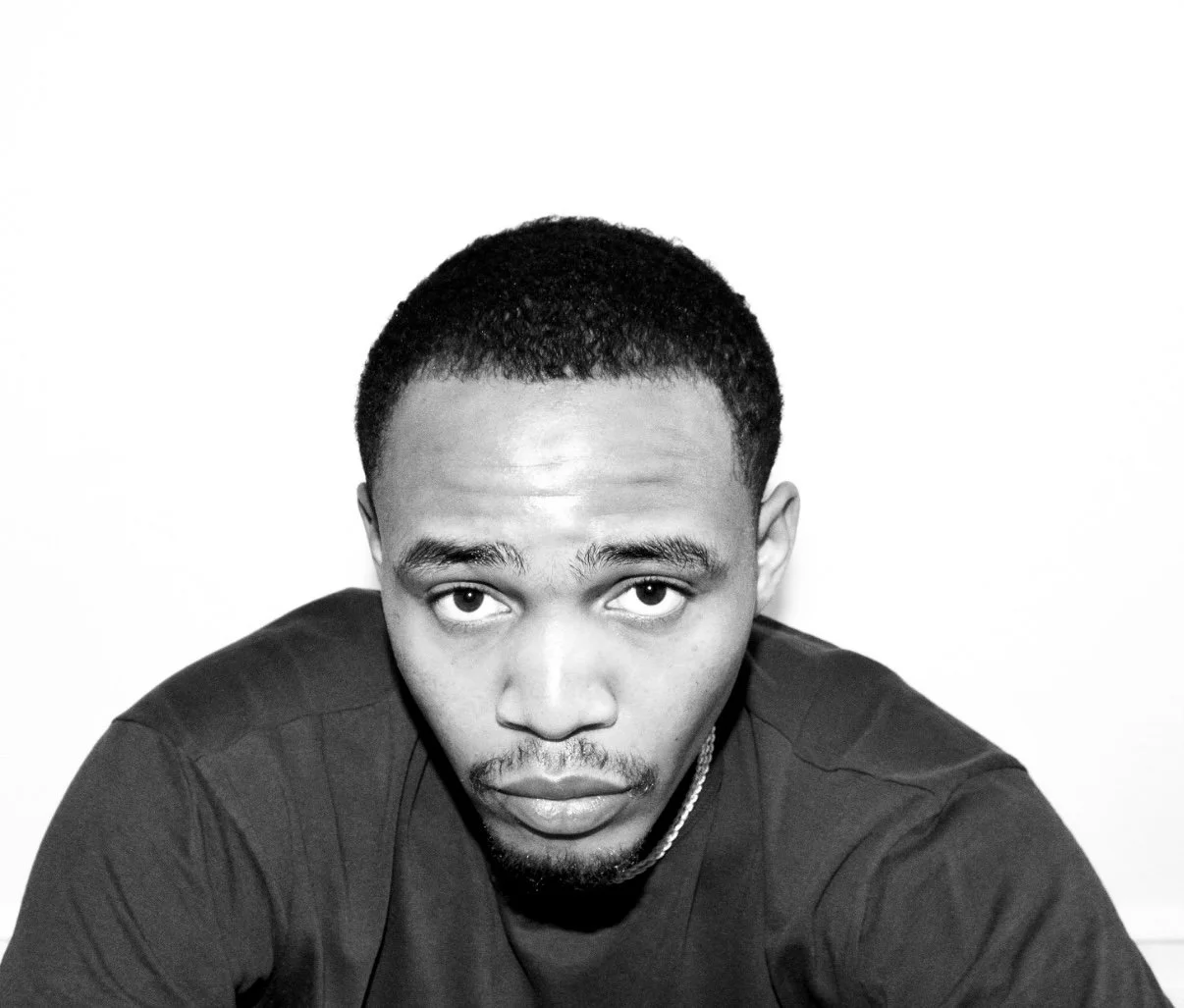 Black and white portrait of a young man with short curly hair, wearing a dark t-shirt. He is looking directly at the camera with a neutral expression, against a plain white background.
