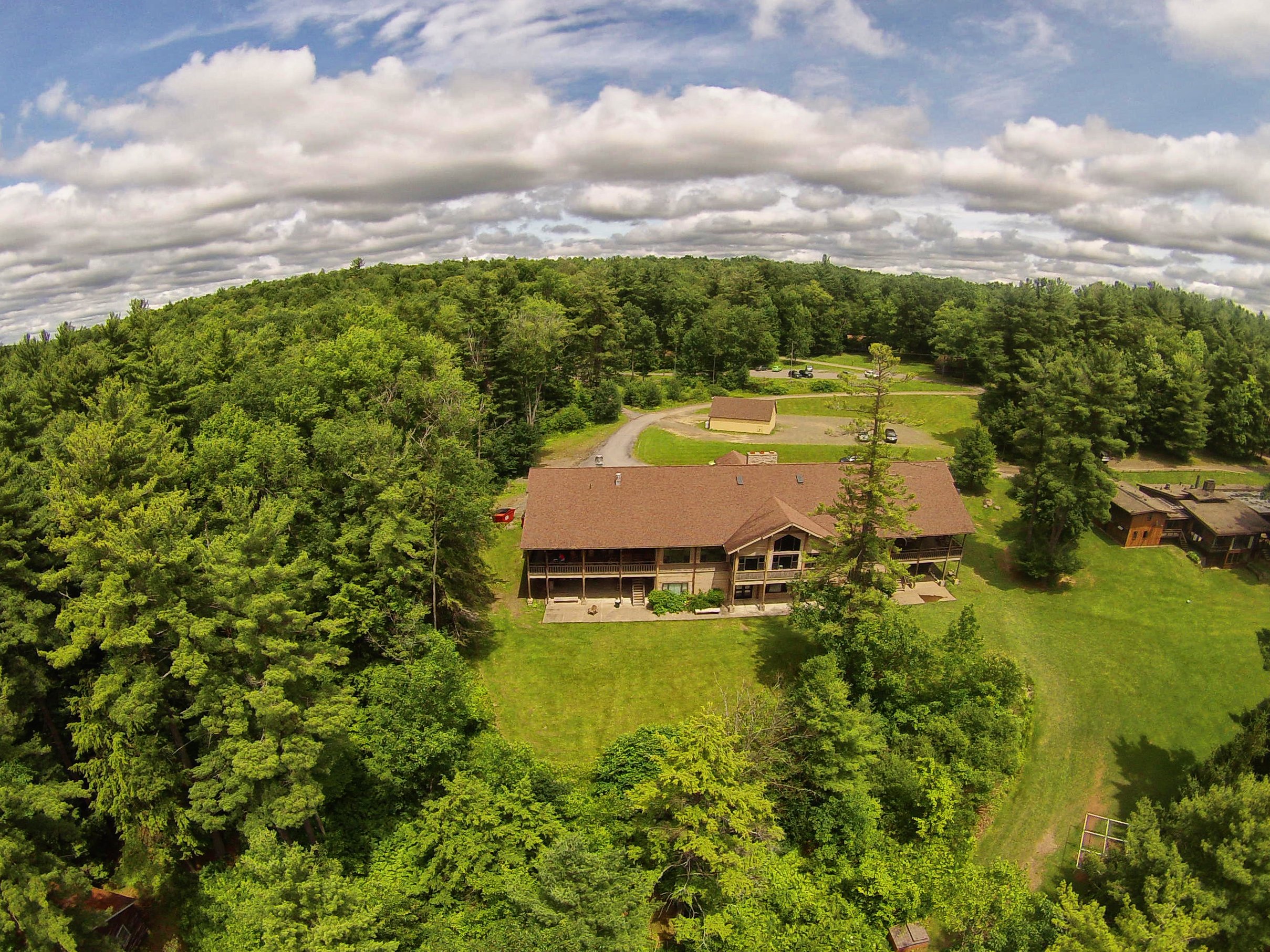 Aerial view of a large house with a brown roof and green lawn surrounded by trees, with a wooded hill in the background and a partly cloudy sky.