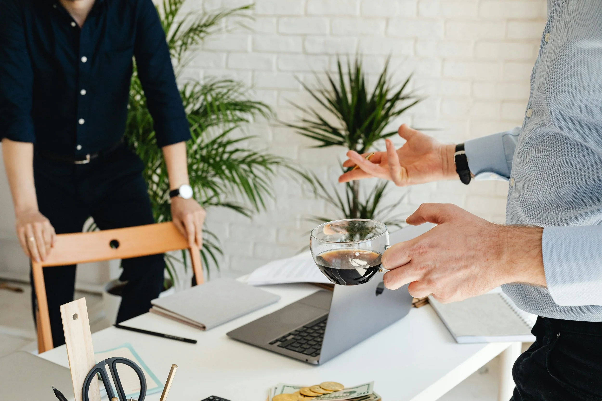 Person holding a glass of red wine in an office setting with a laptop, notebooks, and a potted plant in the background.