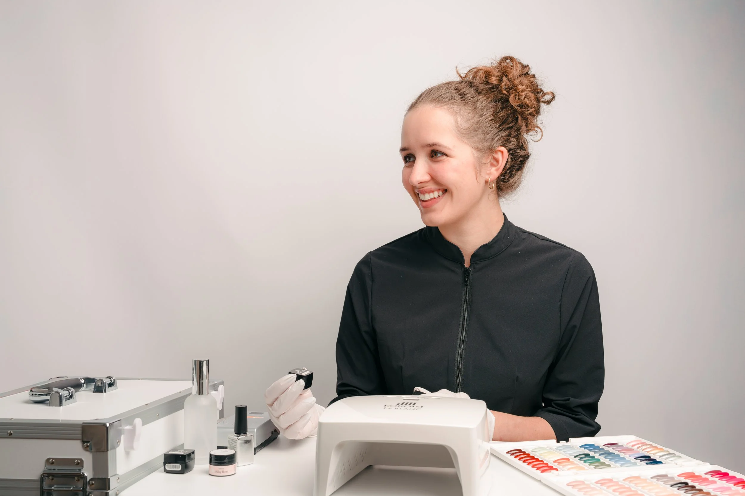 A woman with curly hair, wearing a black zip-up jacket and latex gloves, is smiling while working with nail polish samples and tools at a white table. There are various nail polish colors and a small electronic device on the table, against a plain white background.