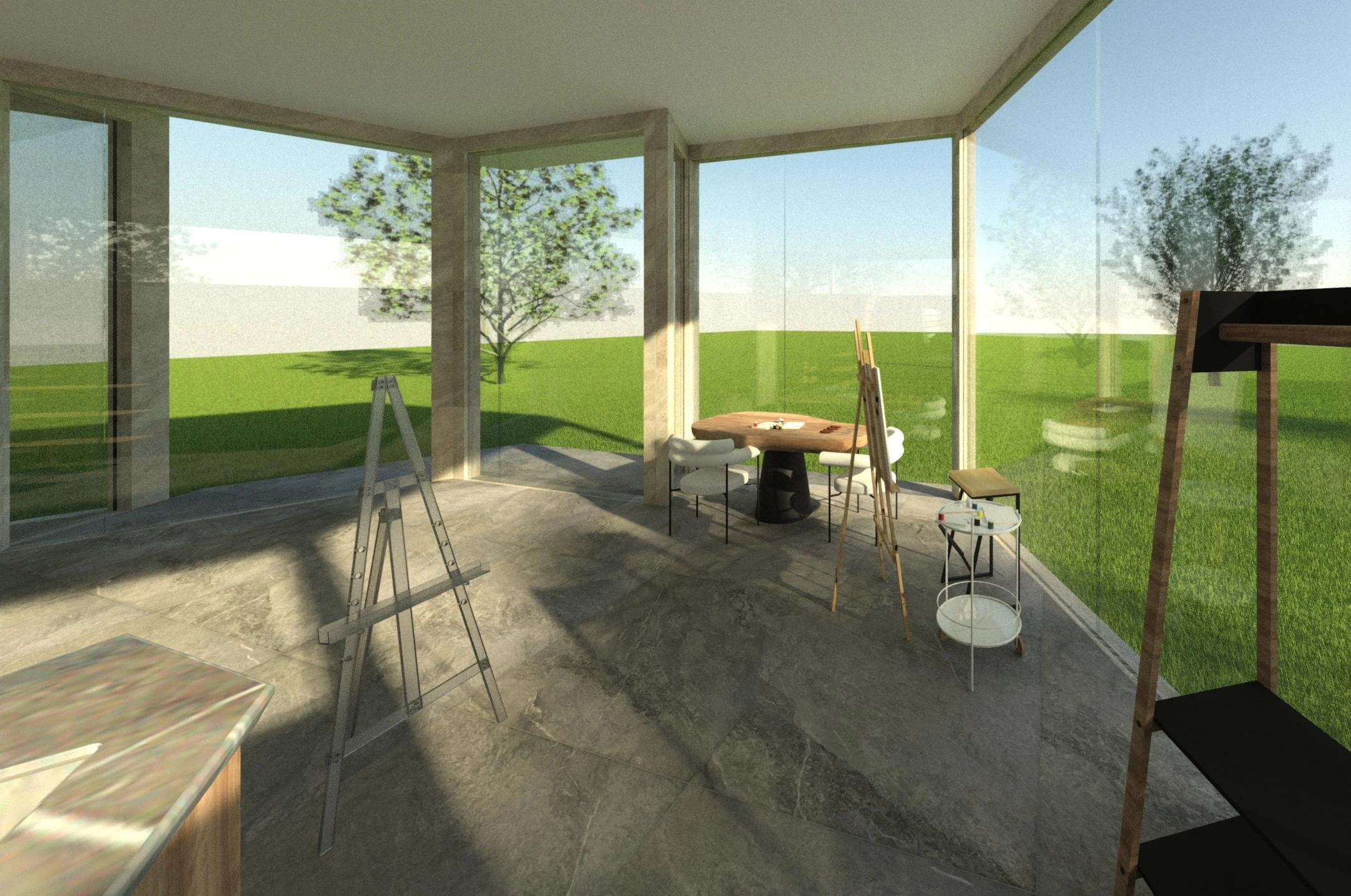 Interior of a sunlit artist's studio with large glass windows showing green grass and trees outside. There are art supplies, tables, and chairs, including an easel and a round table with chairs around it.