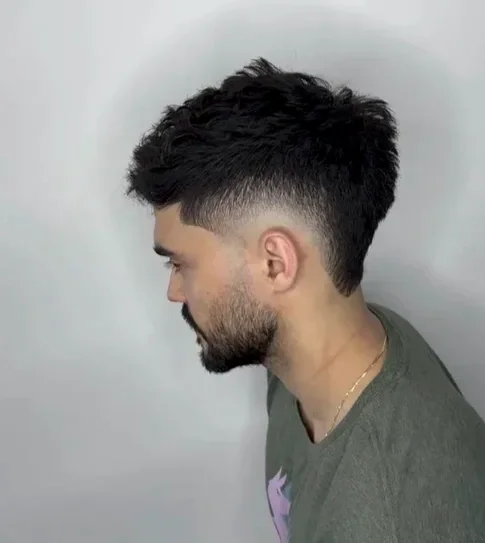 Side profile of a young man with a textured haircut and beard, wearing a gray t-shirt, against a plain light-colored background.