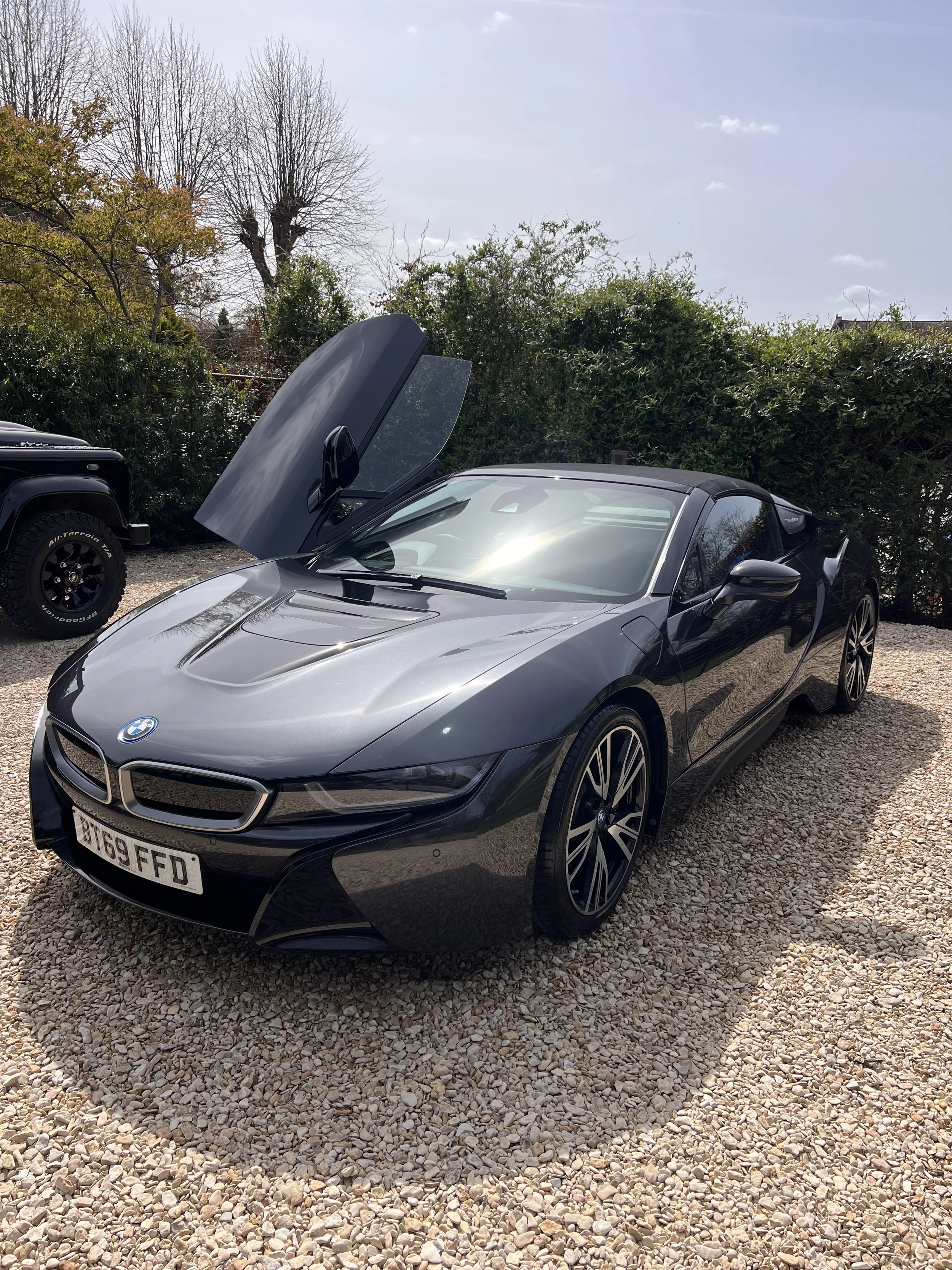 Gray BMW sports car with the front hood and parts of the side visible, parked on a gravel surface with green bushes and trees in the background.