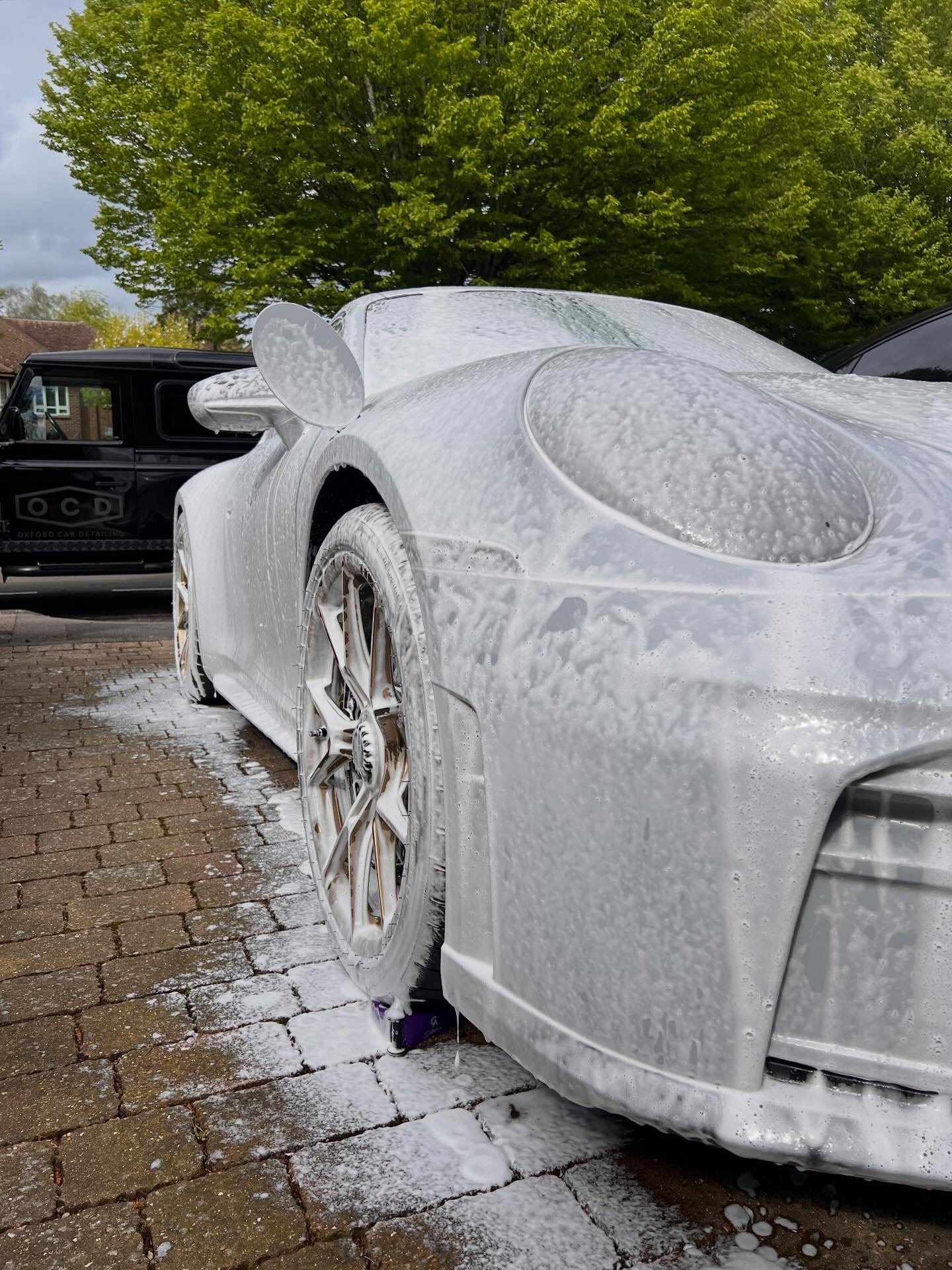 White sports car covered in soap suds during a car wash, with a black vehicle and a large green tree in the background.