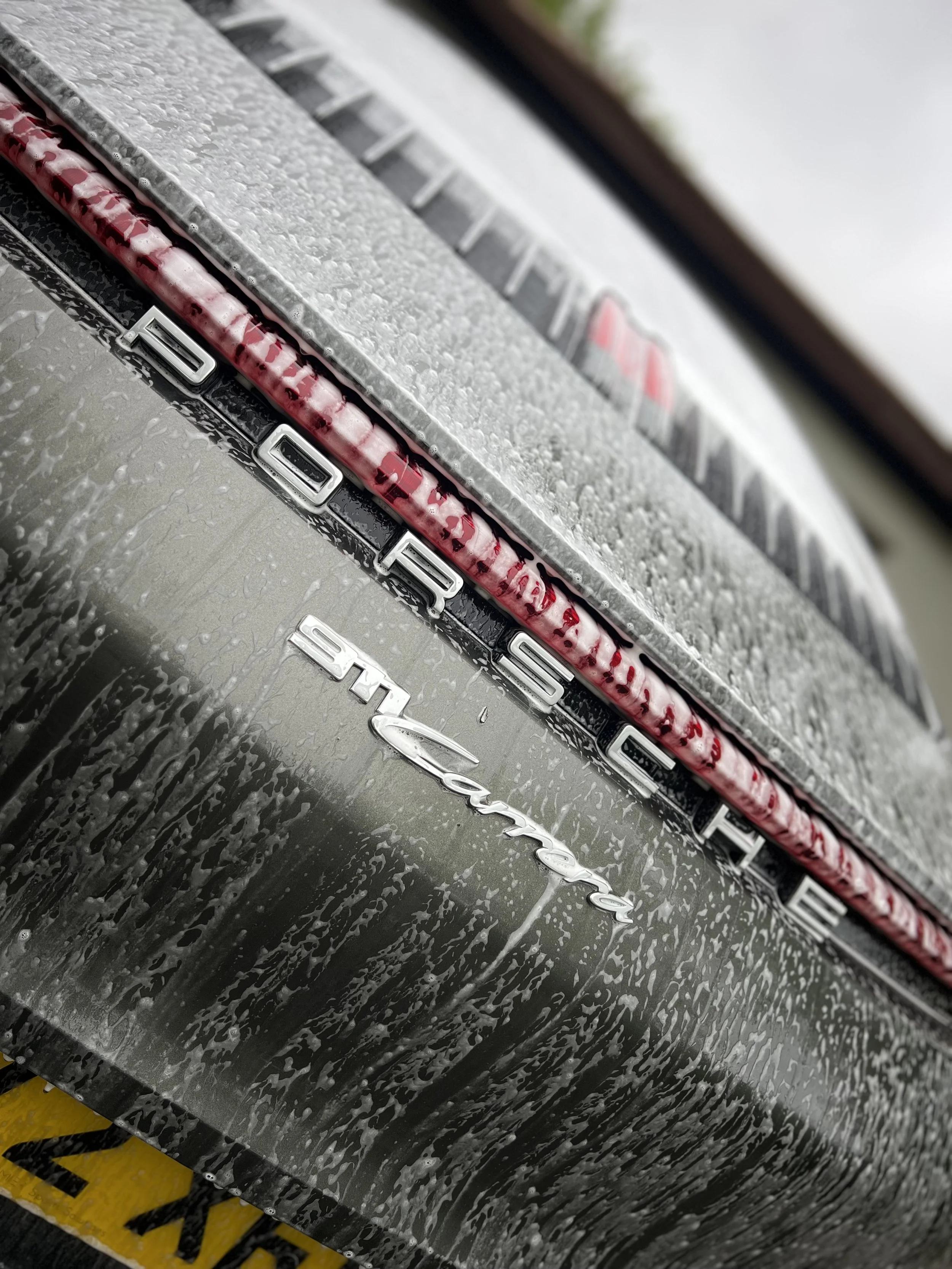 Close-up of the rear part of a black Porsche vehicle showing the model badge and water droplets after washing.