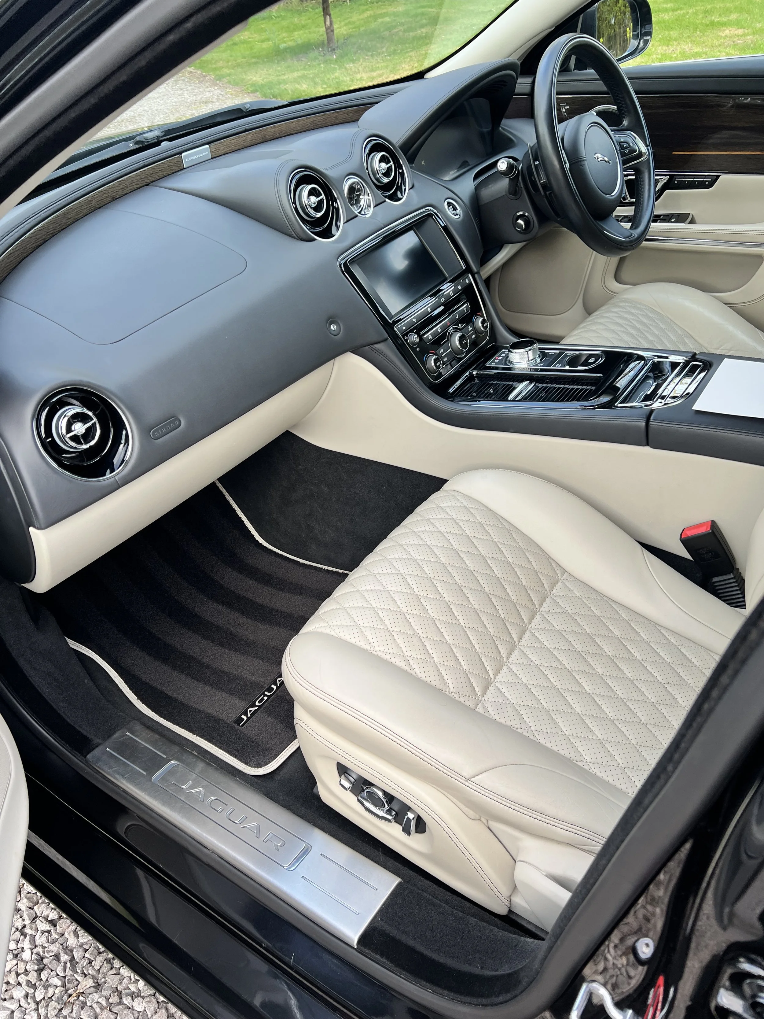 Interior of a luxury black Jaguar car showing the dashboard, steering wheel, and beige quilted leather driver seat.