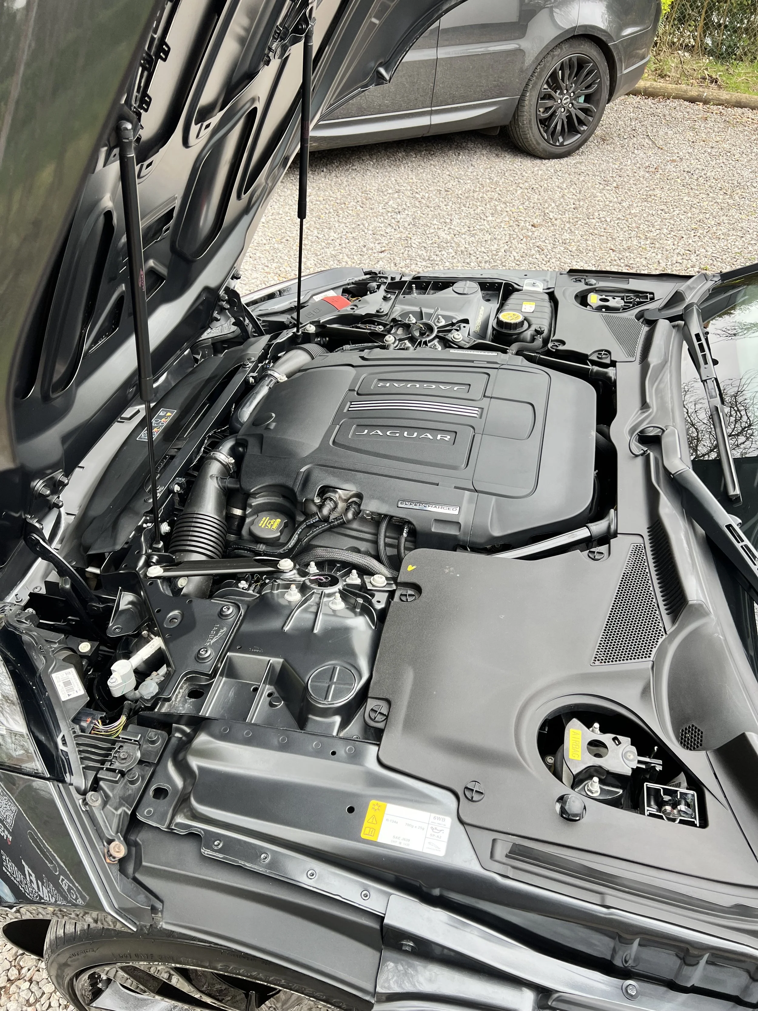 Car with open hood showing engine compartment. The engine cover displays the Jaguar logo and indicates a supercharged engine.