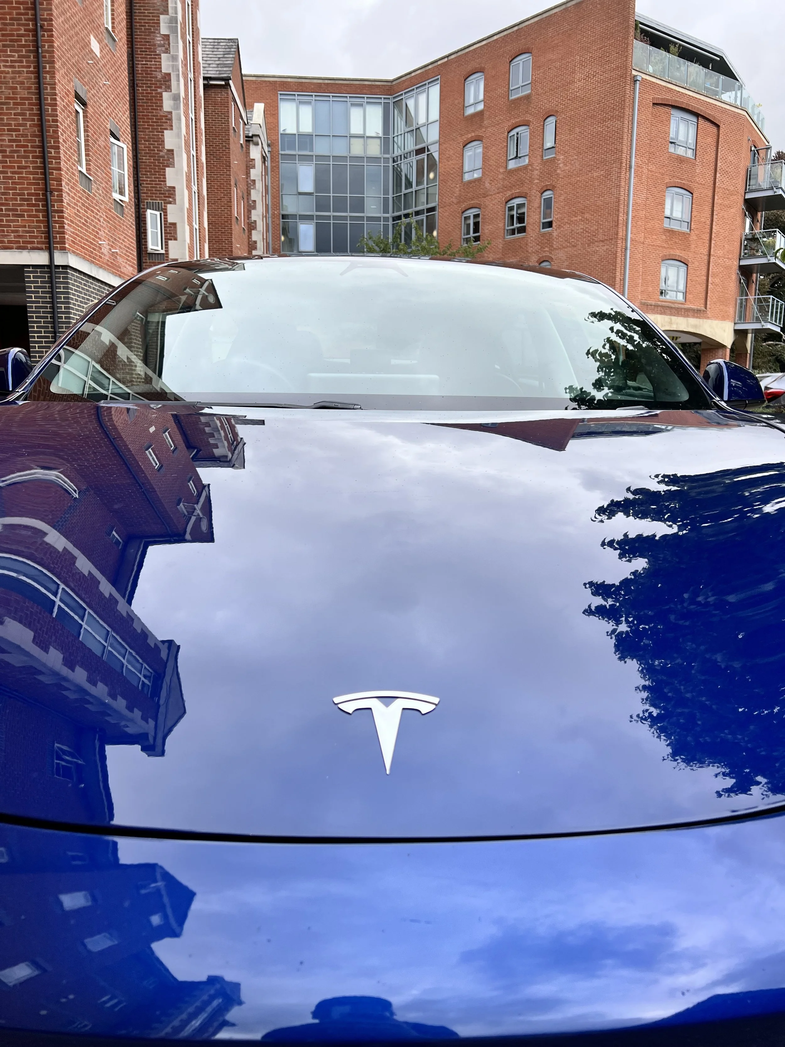Front view of a dark blue Tesla car parked outdoors after full valet and detailing. The car's hood reflects the buildings and cloudy sky.