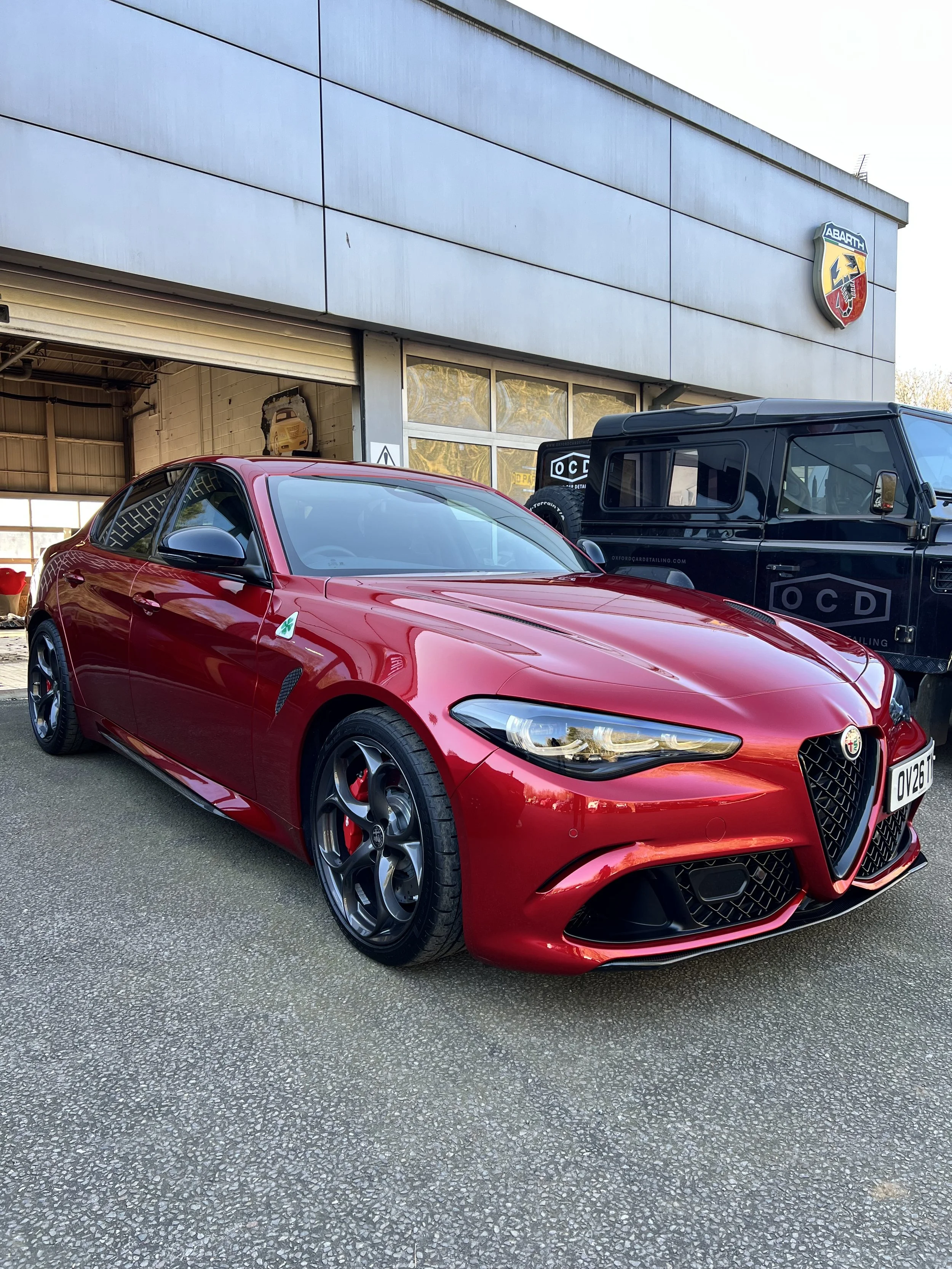 Red sports car parked in front of a garage with a partially open door, beside a black van, outside a building with a sign that reads 'Abarth' and an emblem above.
