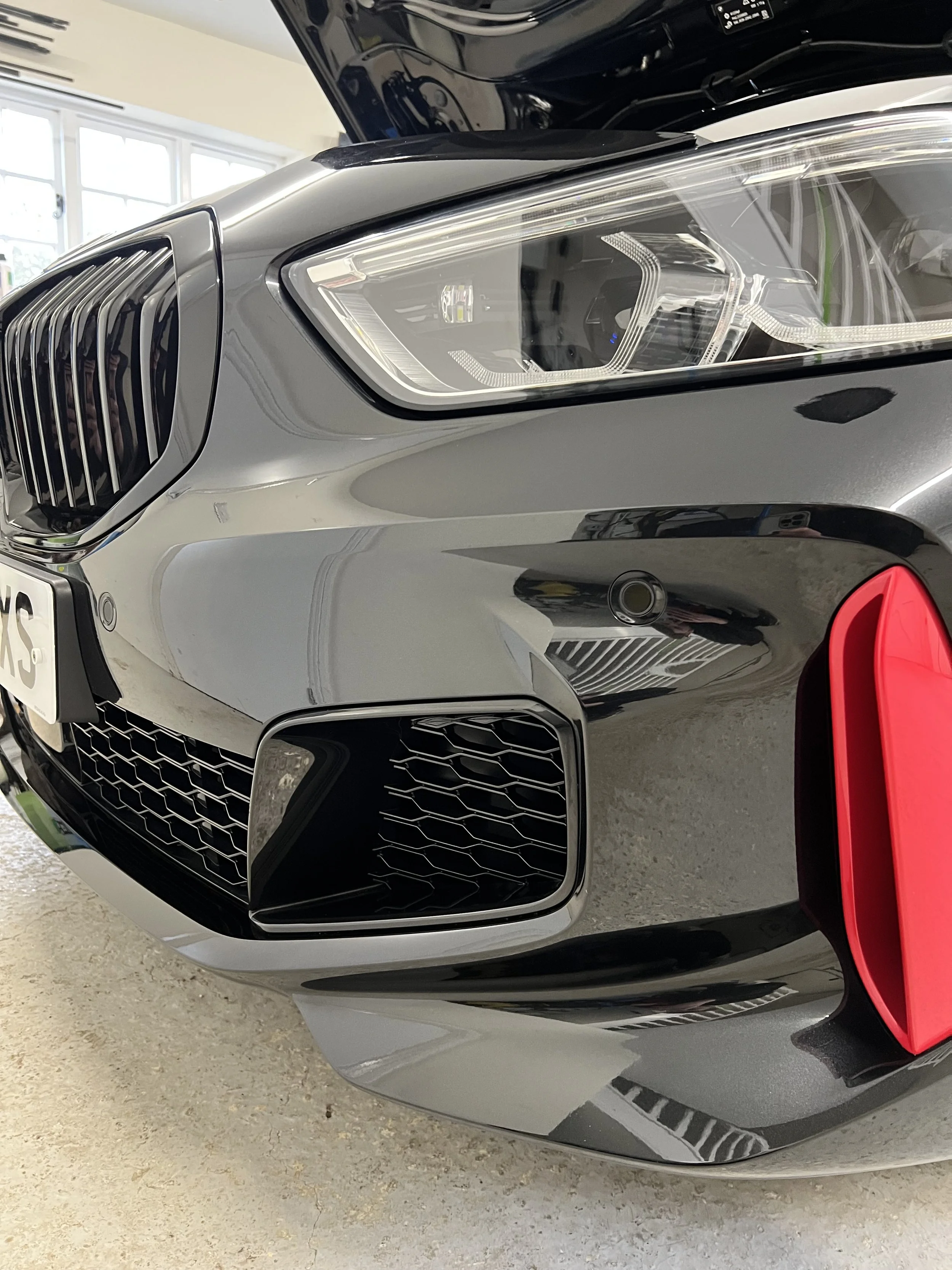 Close-up of the front left side of a silver luxury car, showing the headlight, grille, and part of the bumper with a red accent near the wheel.