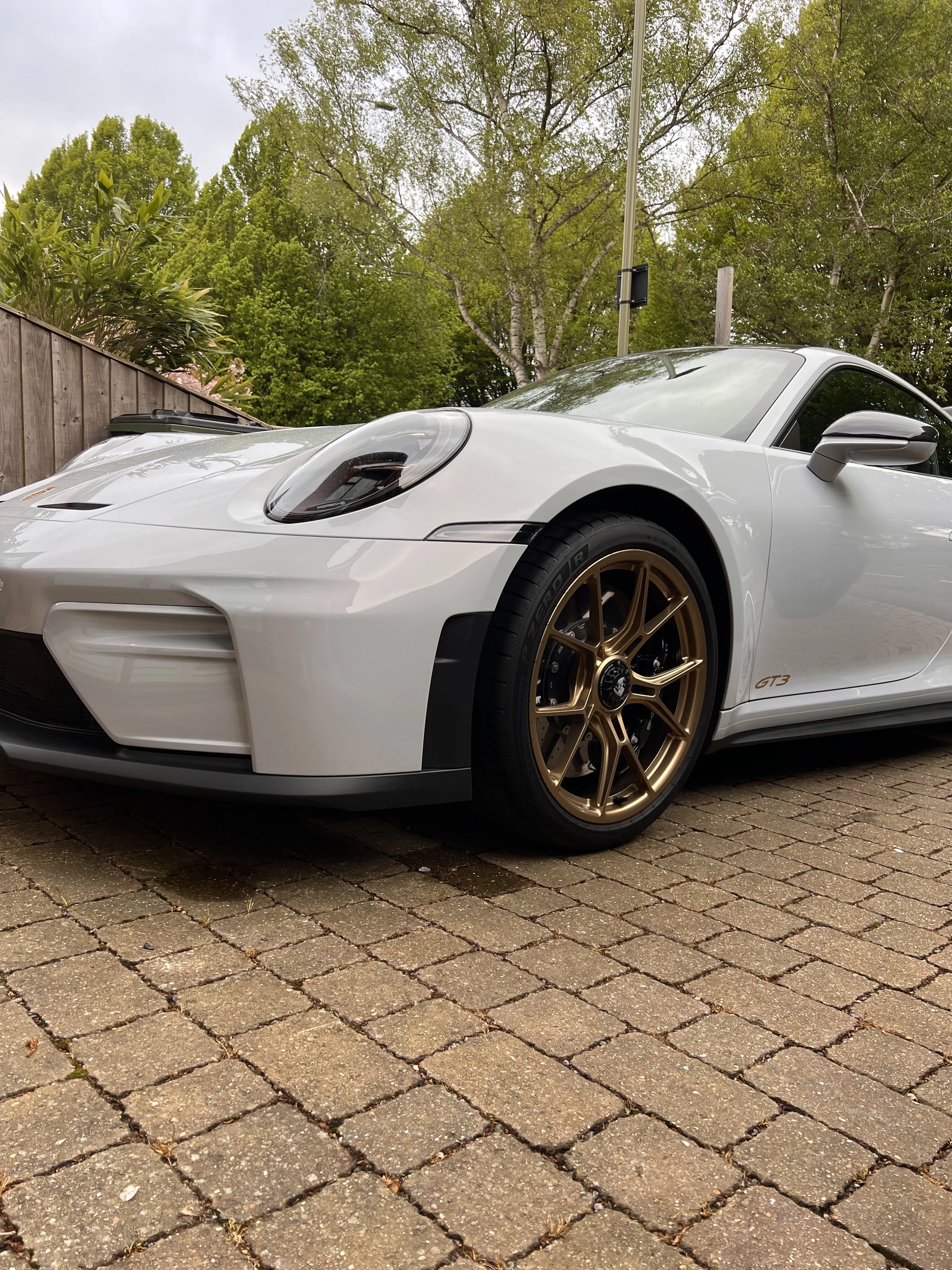 A white Porsche GT3 sports car with gold wheels parked on a brick driveway surrounded by green trees and a wooden fence.