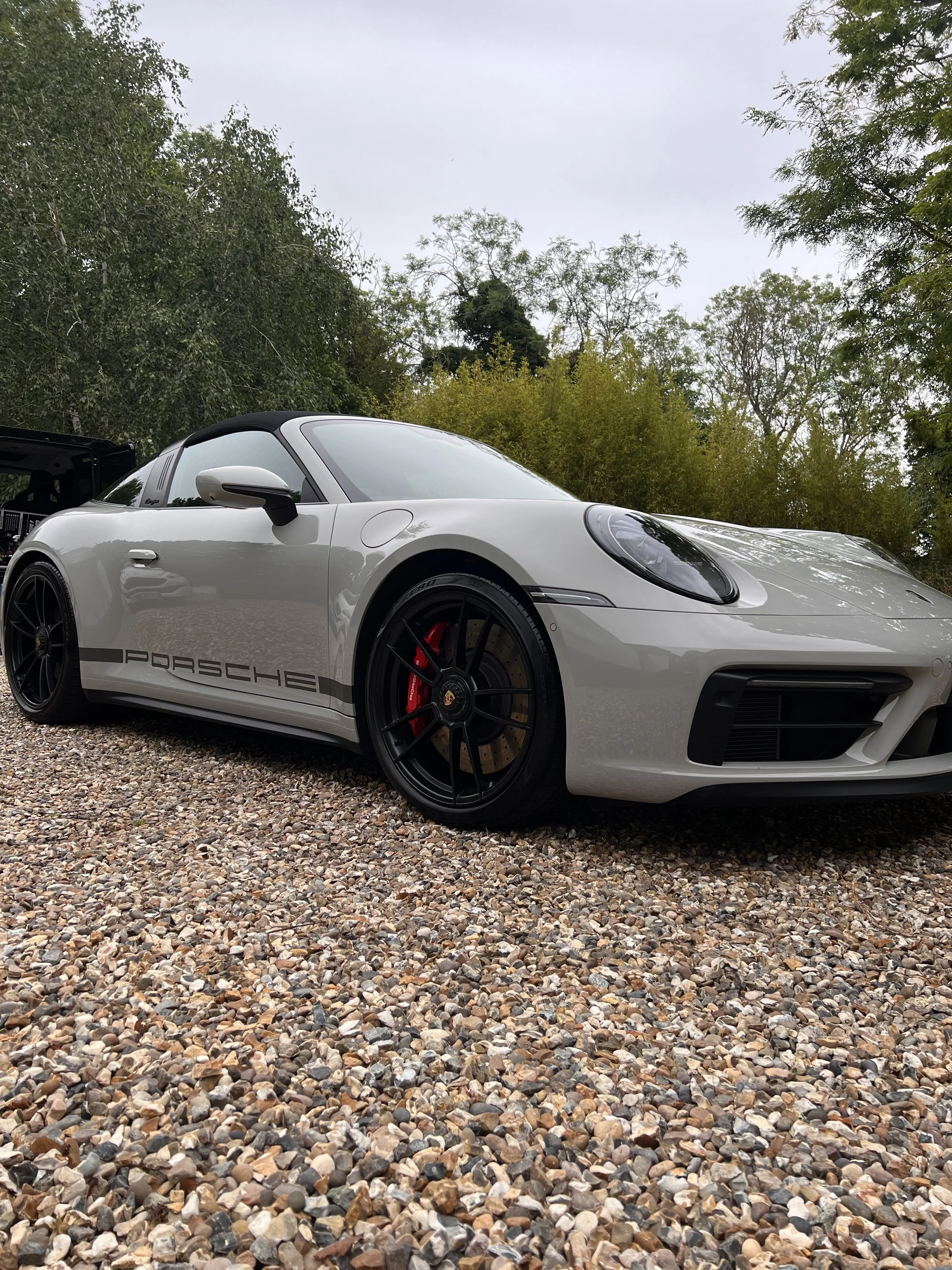 A white Porsche sports car with black accents parked on a gravel surface among trees under an overcast sky.