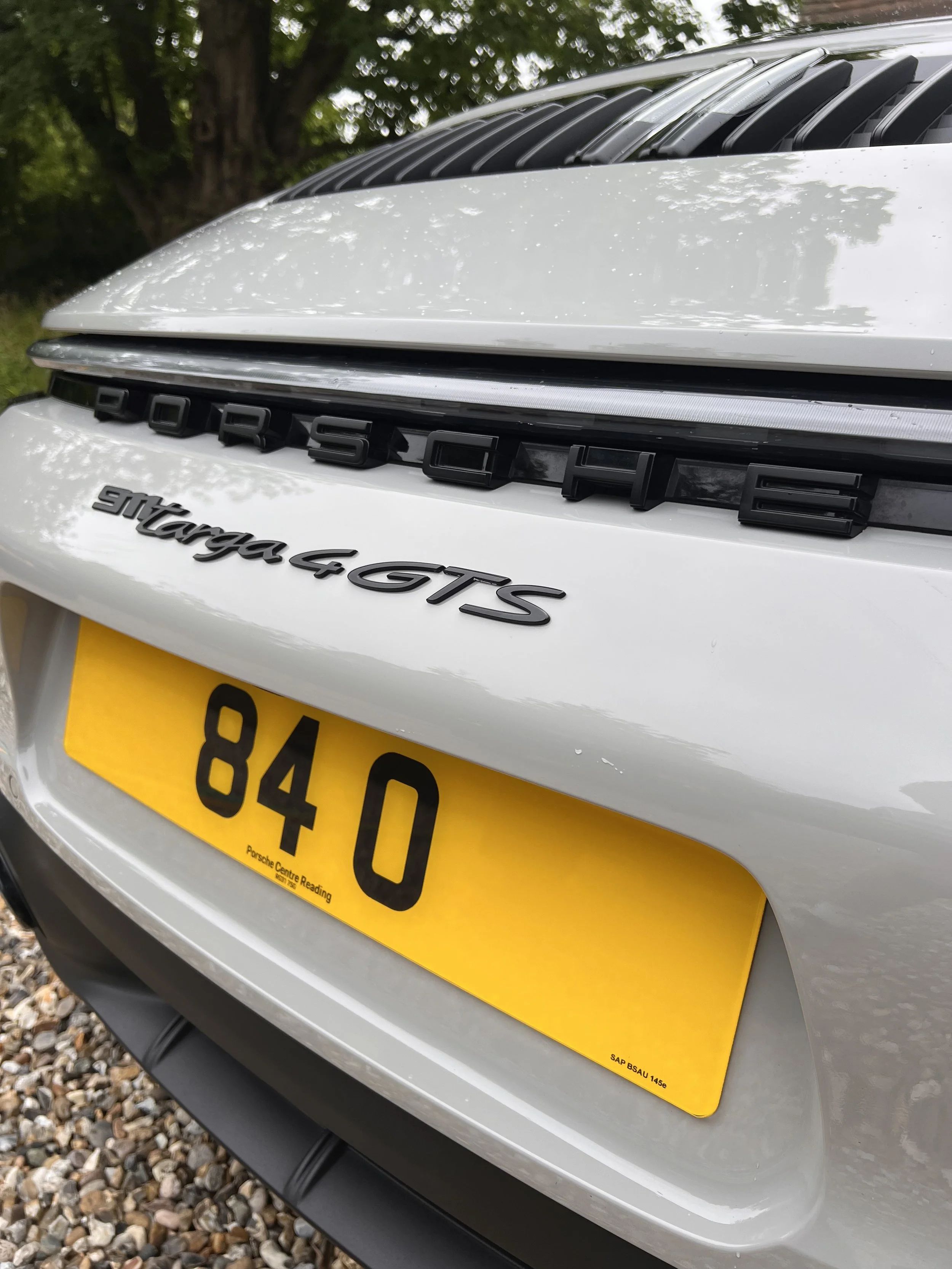Close-up of the rear of a white Porsche vehicle showing the model badge 'Stuttgart 4 GTS' and a yellow UK license plate with the number '84 8O', with a tree and gravel ground in the background.