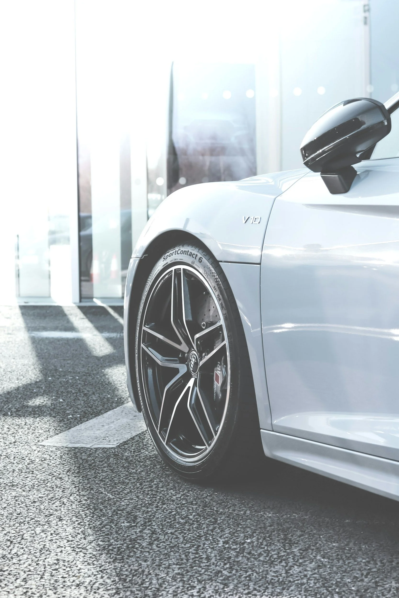 Close-up of the front part of a silver luxury sports car with black custom wheels, parked outside near a building with glass doors, sunlight reflecting off the car surface.