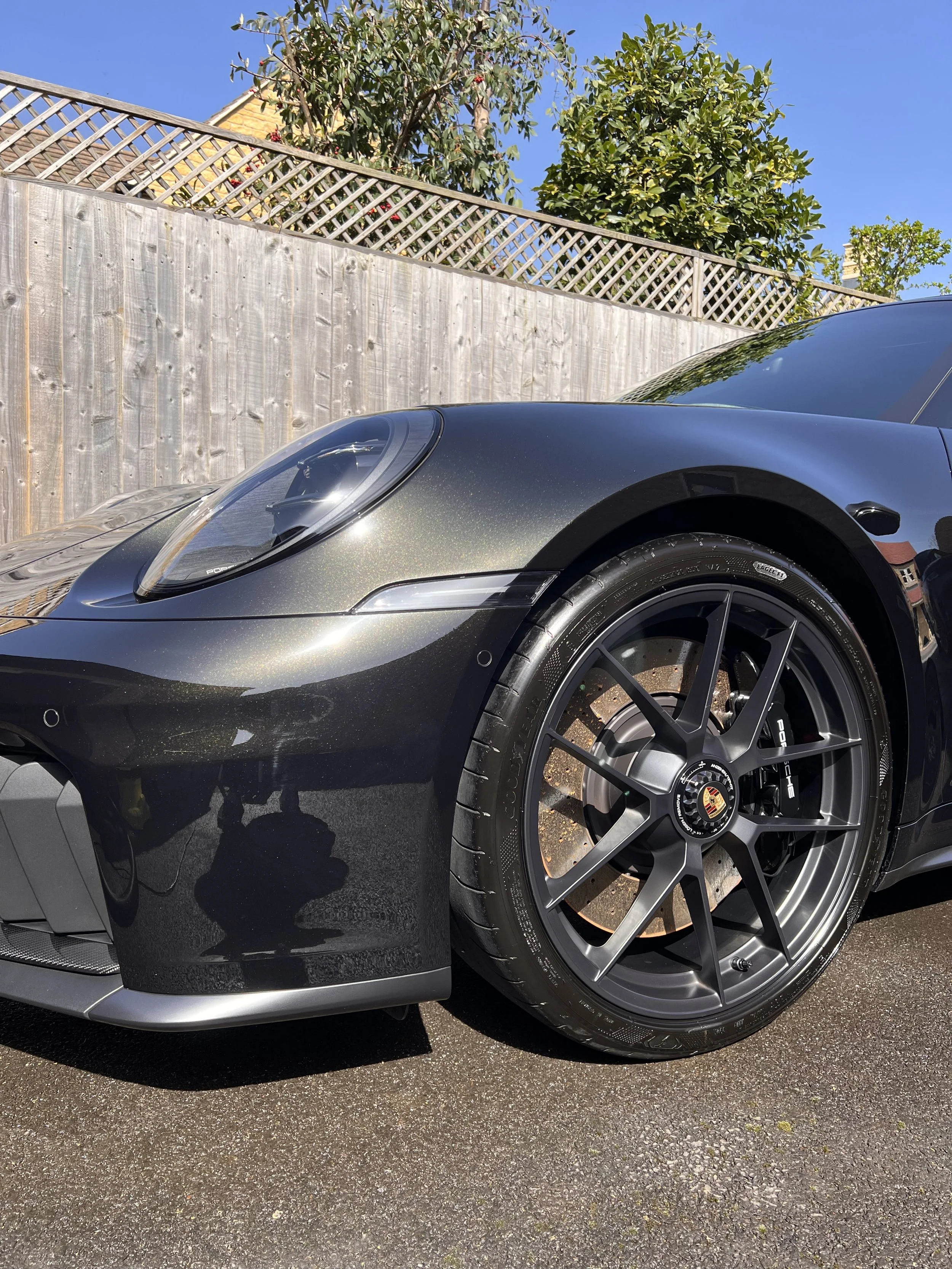 Close-up of the front part of a dark gray Porsche sports car parked on a street, focusing on the wheel, headlight, and fender, with a wooden fence, green bushes, and blue sky in the background.