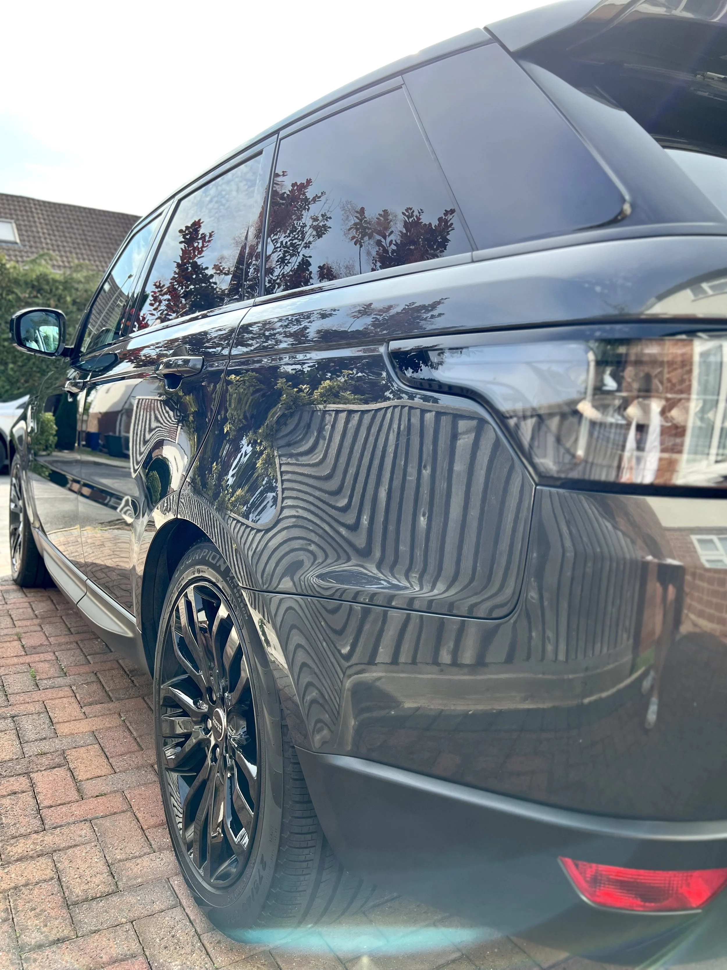 Black SUV parked on a brick driveway with reflections of trees and houses on its shiny surface.