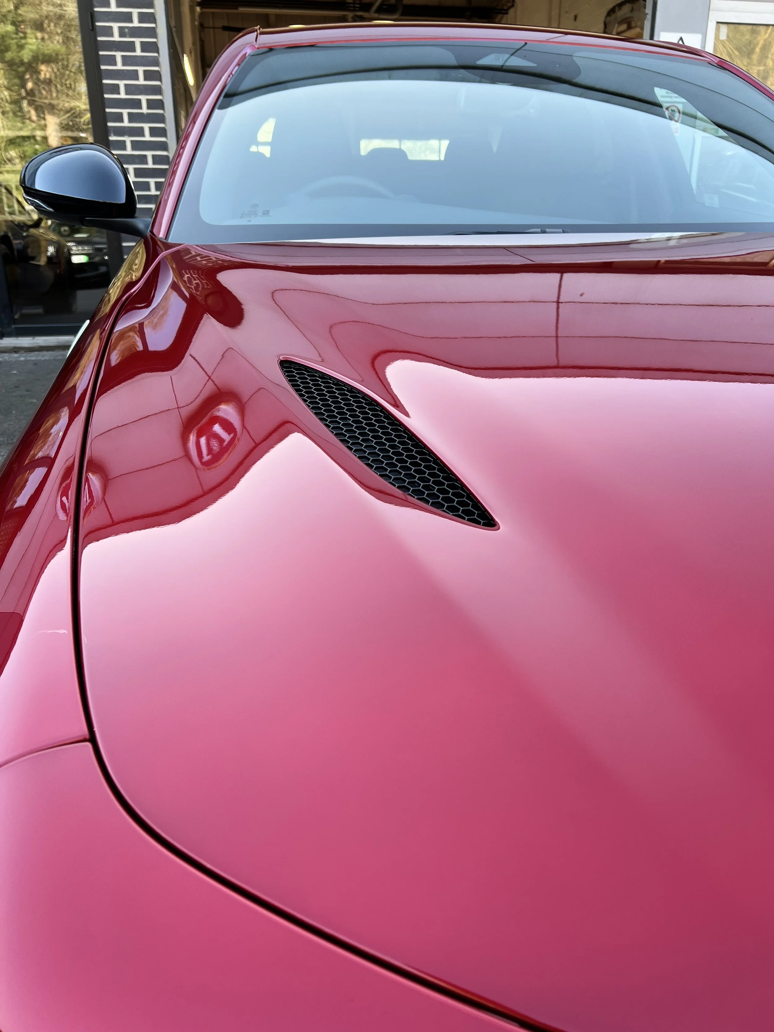 Close-up of a red sports car's front corner, showing the hood, side mirror, and windshield.
