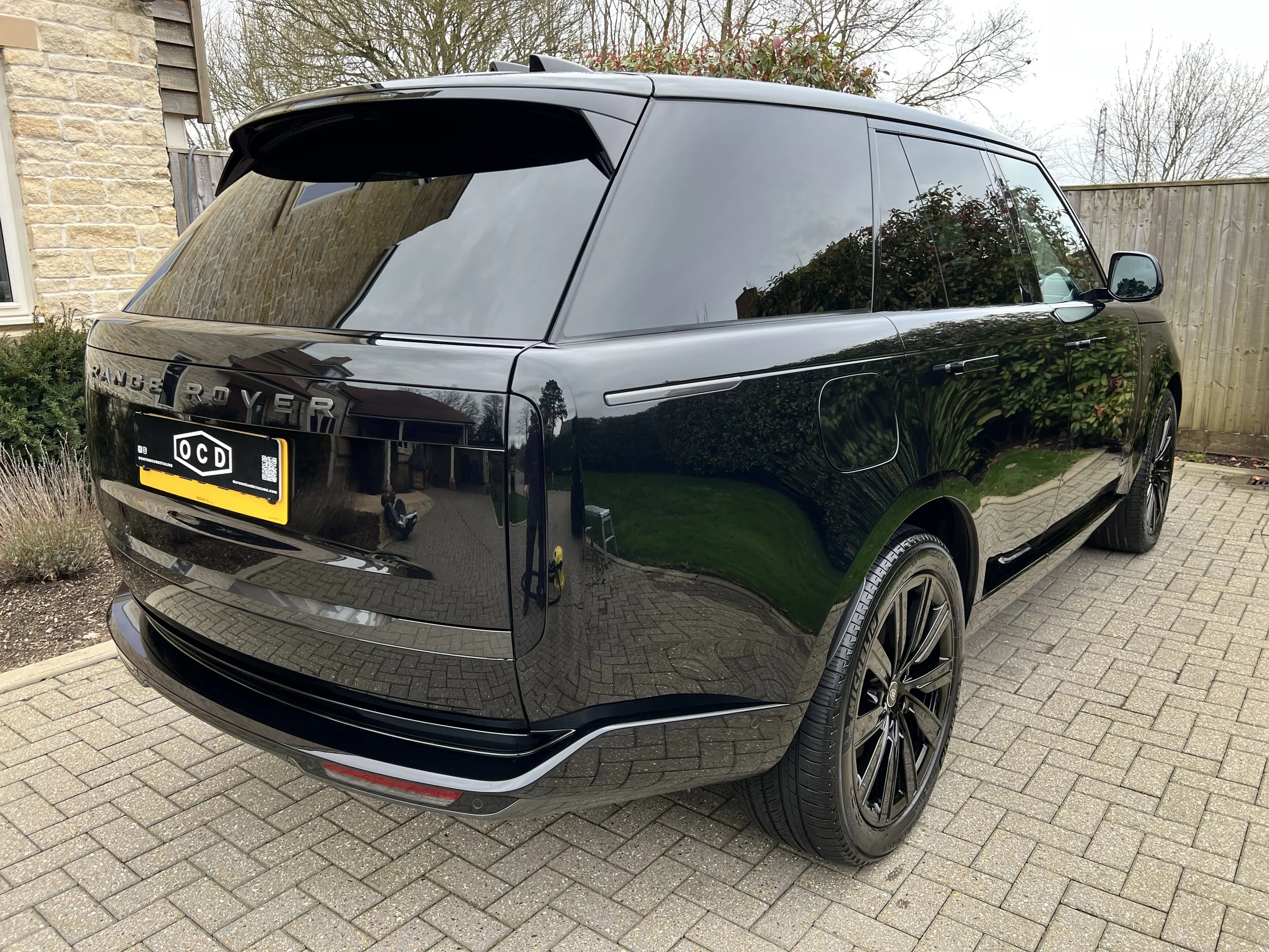 A black Range Rover SUV parked on a brick driveway near a house, with trees and a wooden fence in the background.