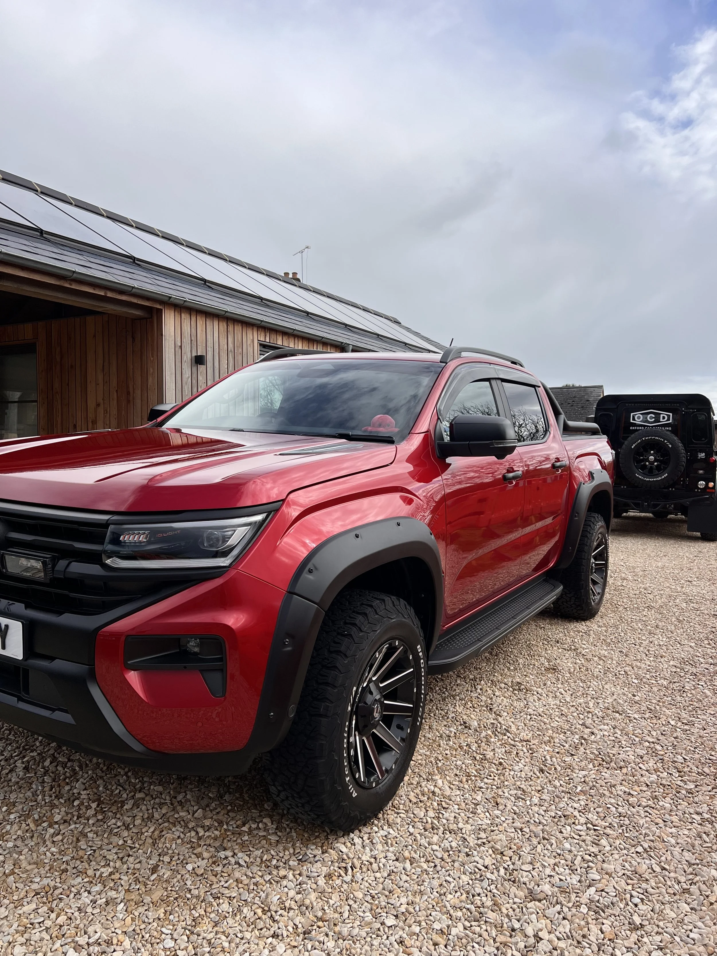 Red off-road pickup truck parked on a gravel driveway next to a wooden house with solar panels on the roof, cloudy sky in the background.