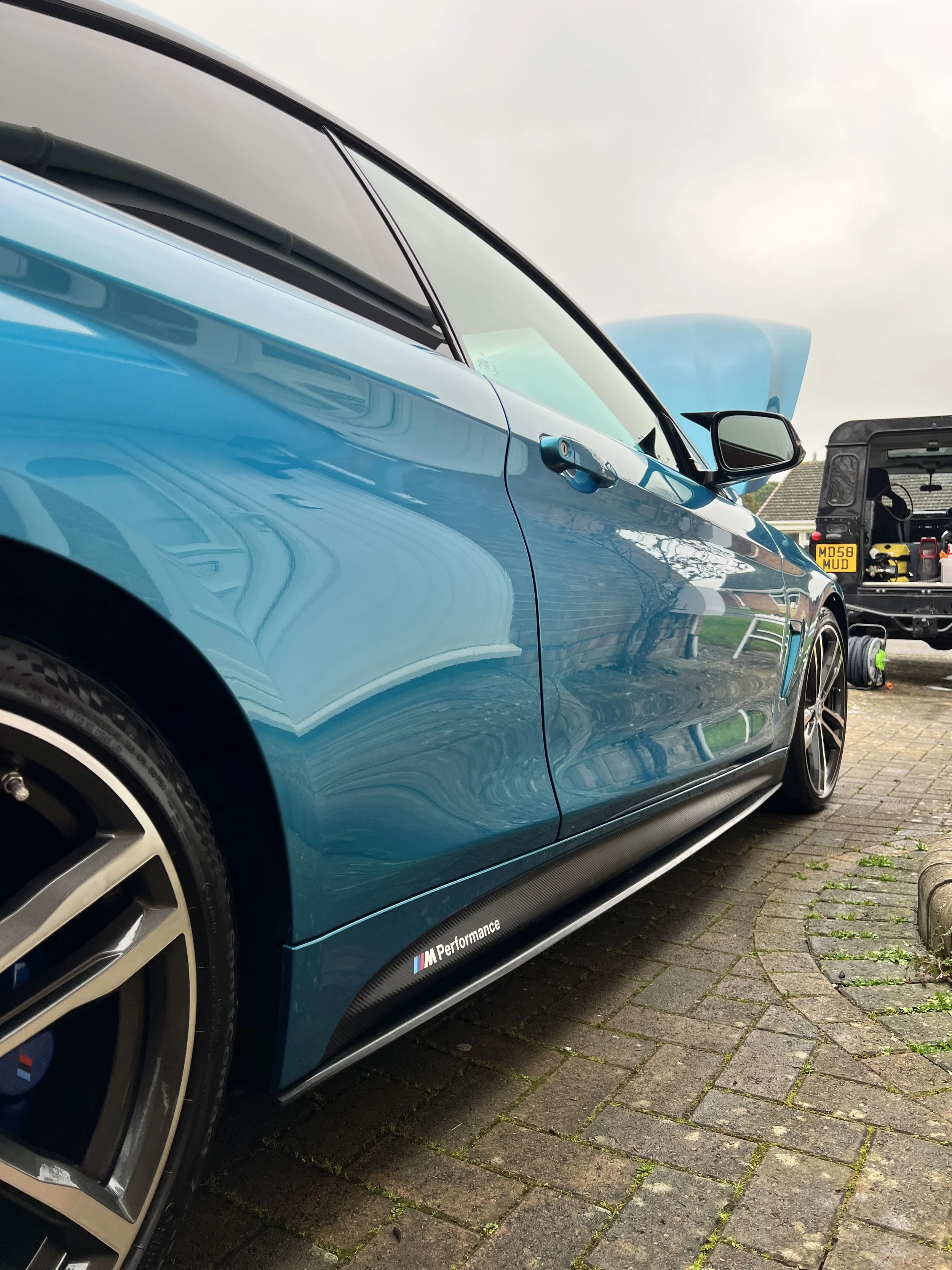A blue BMW M Performance car parked on a brick driveway, with a black van and some tools in the background under an overcast sky.