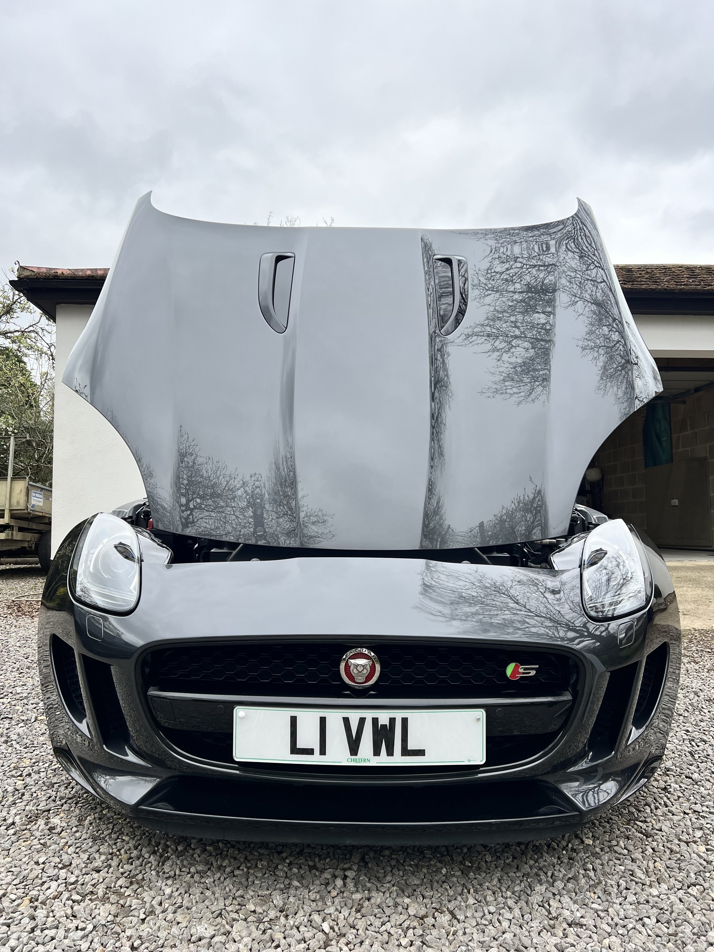 Front view of a gray Jaguar car with its hood open, parked on a gravel driveway in front of a white house with a brick wall and garage. The car's license plate reads 'LIVWL'.
