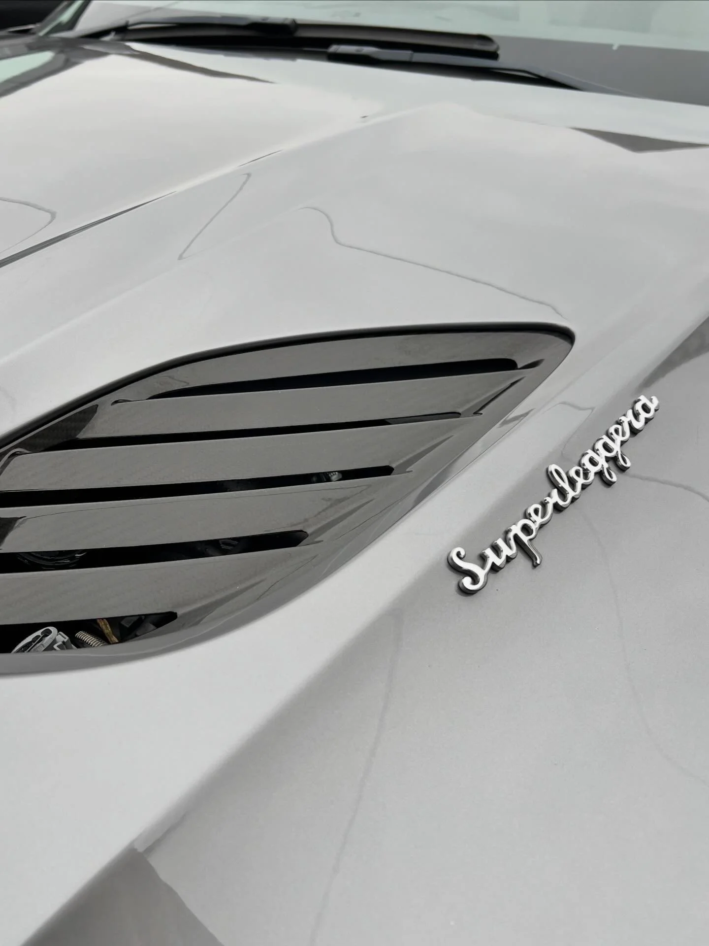Close-up of a white car with a silver 'Superleggera' badge on the side, featuring air vents on the front fender.