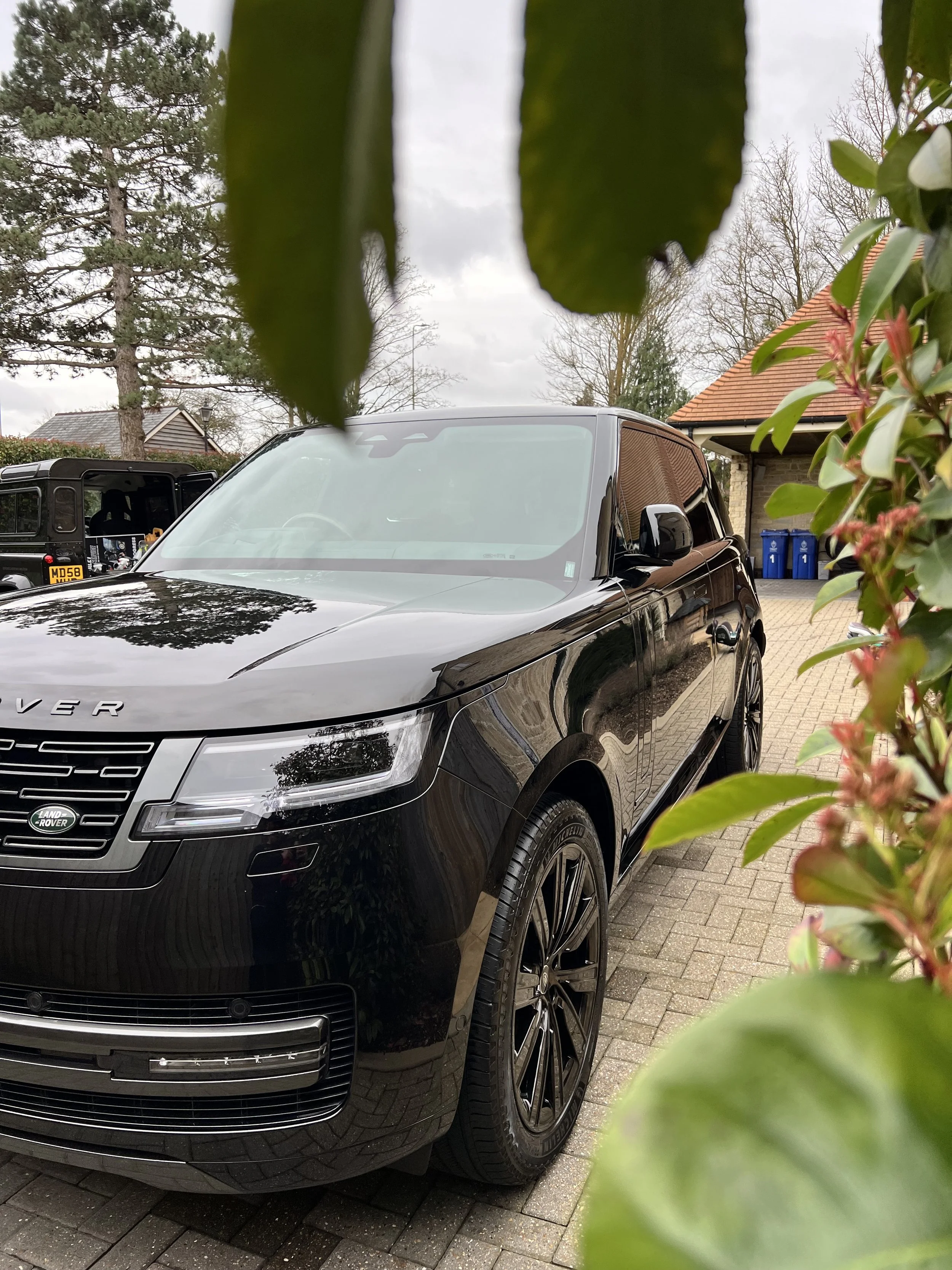 A black Land Rover SUV parked on a brick driveway, partially obscured by green leaves in the foreground. In the background, a house with a pitched roof and several blue recycling bins are visible.