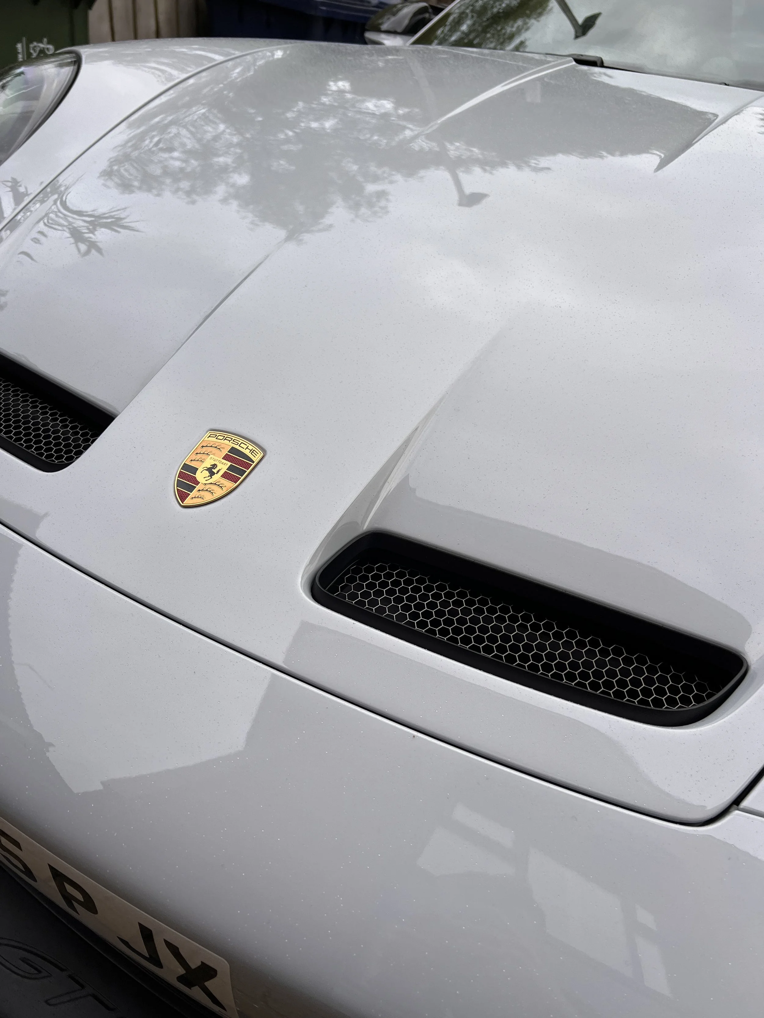Close-up photo of the hood of a silver Porsche sports car, displaying the Porsche badge and mesh air vents.